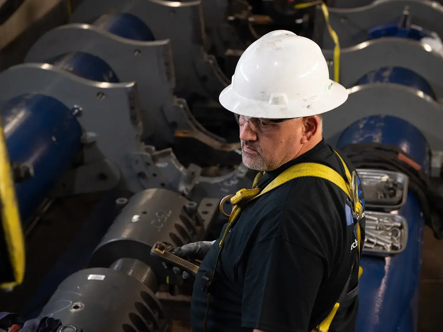 A worker wearing a white safety helmet, safety glasses, and a black shirt with yellow safety harness inspects mechanical components in an industrial setting filled with large blue metal machinery parts.