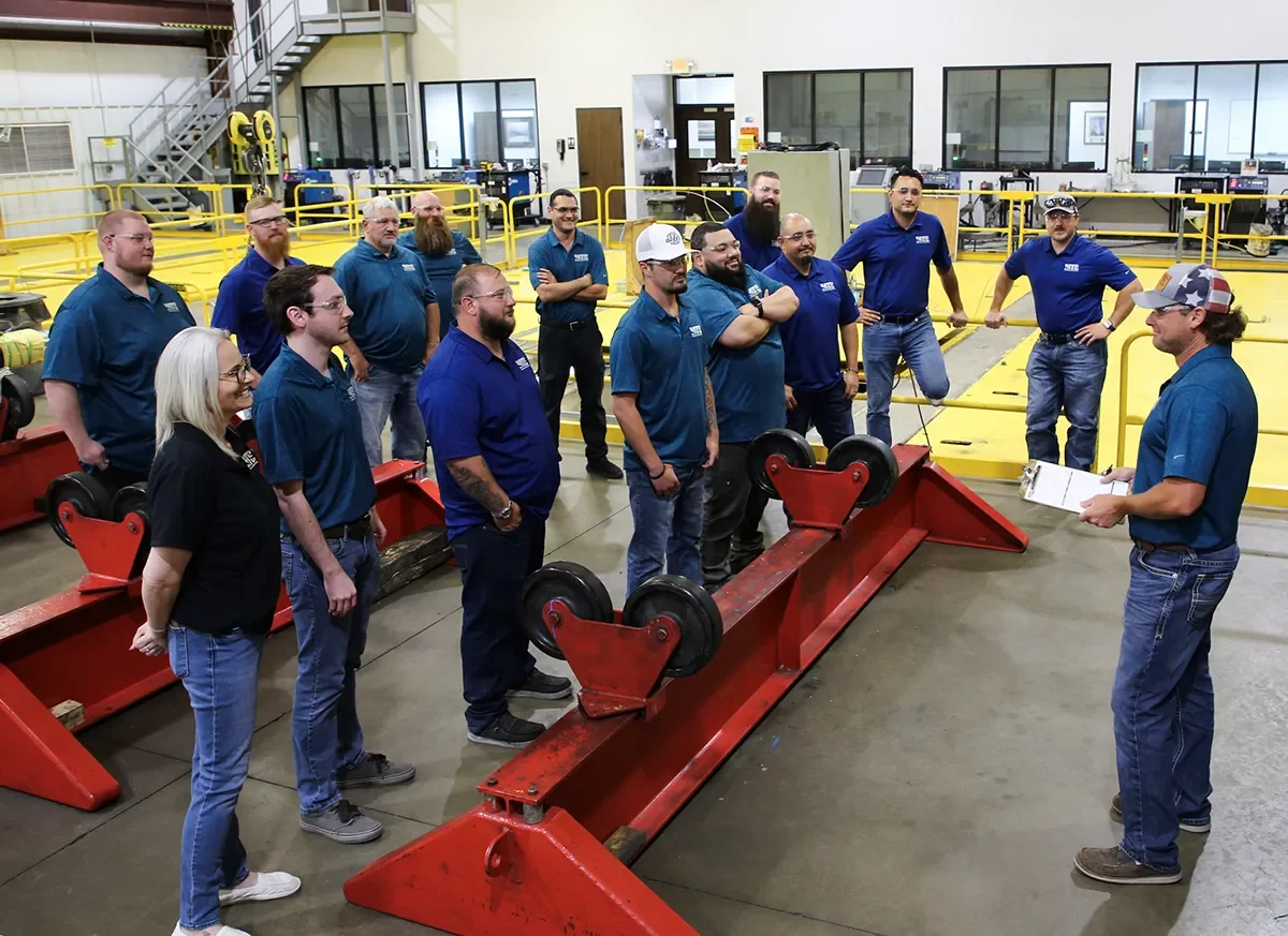A group of MTS team members standing in a factory or warehouse, listening to a person holding a clipboard, with industrial equipment and safety barriers in the background.