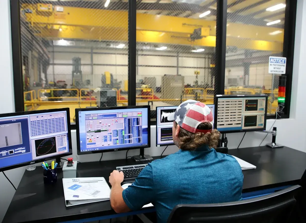 A control room with a person operating multiple monitors showing data, with a view of an industrial workshop or manufacturing facility through large windows in the background.