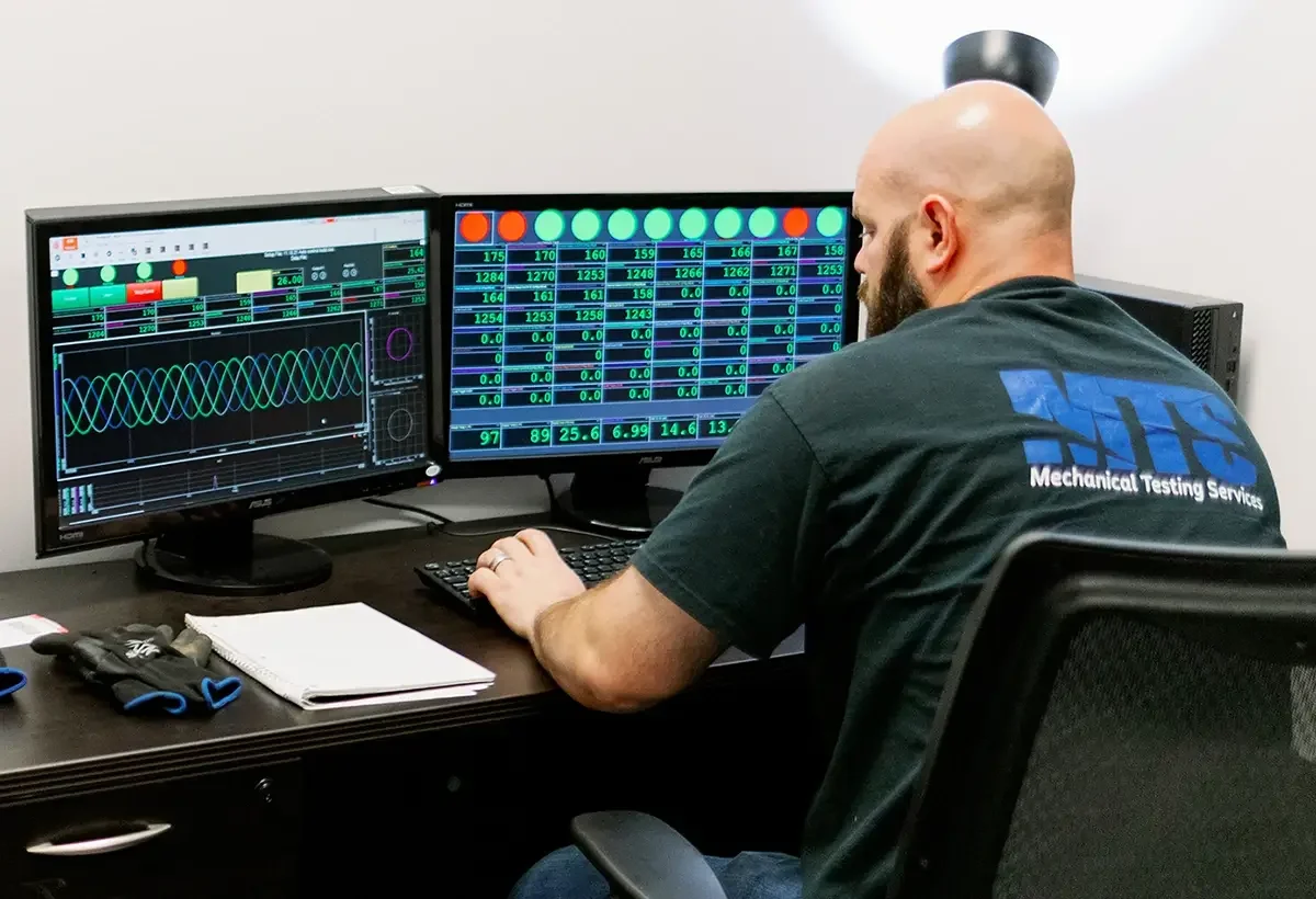 A man with a shaved head and beard using dual computer monitors displaying data related to mechanical testing, sitting at a desk with notebook and work gloves.