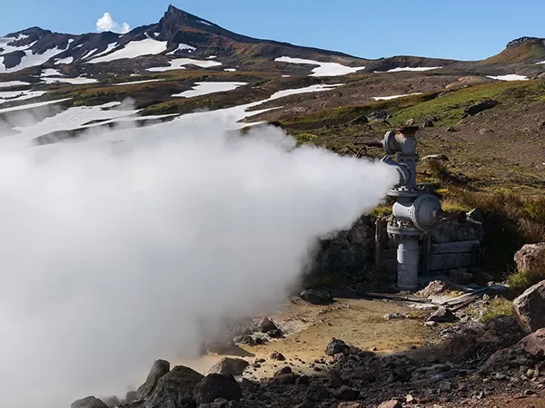A geothermal steam vent releasing steam in a mountainous landscape with patches of snow and rocky terrain.