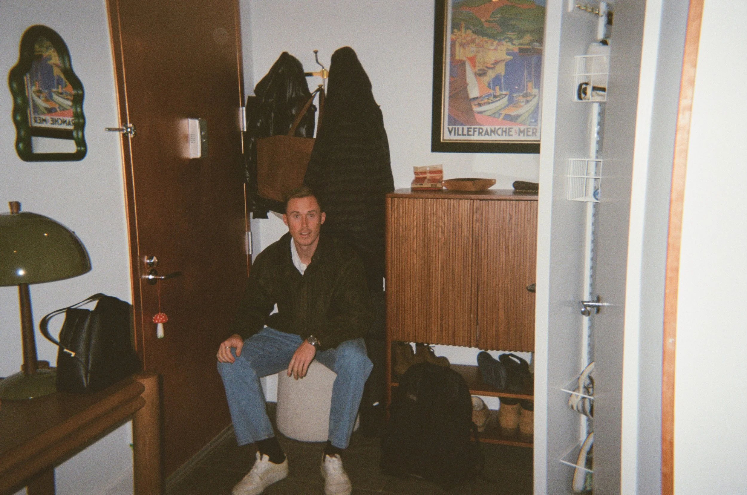 A man sitting on a white stool in an entryway, surrounded by coats, bags, and shoes, with a framed colorful poster of Villefranche-sur-Mer on the wall.