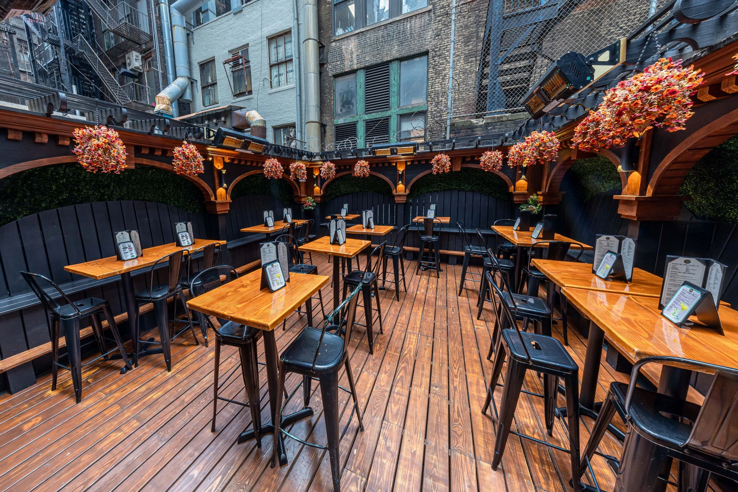 Empty outdoor restaurant patio with wooden tables and black chairs, decorated with hanging flower arrangements, surrounded by black walls and buildings with fire escape stairs.