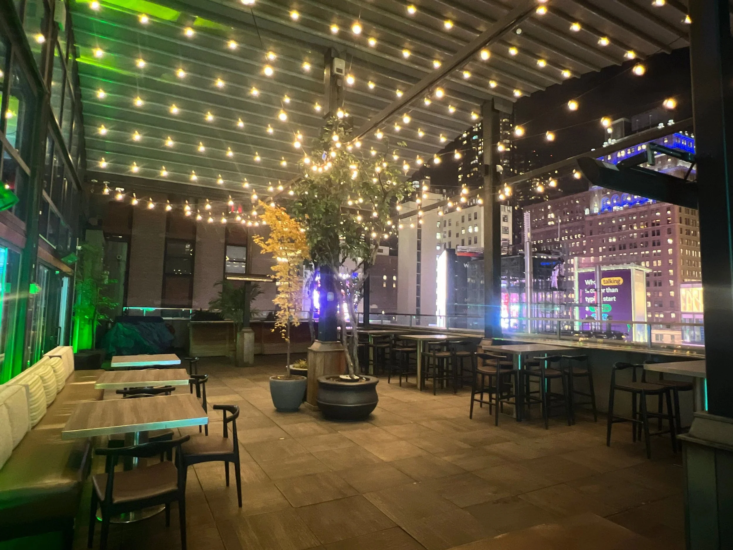 Nighttime rooftop bar with string lights hanging from the ceiling, featuring tables, chairs, and potted plants, overlooking city skyscrapers and billboards.