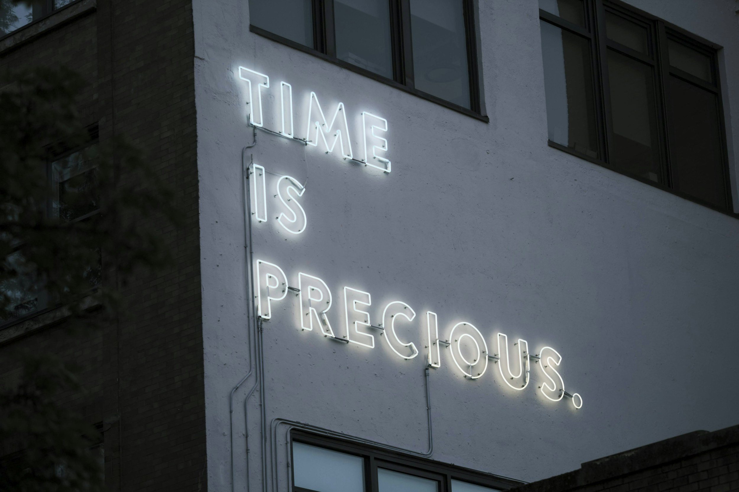 Neon sign on a white building wall that reads 'Time is Precious' in light blue and white neon lights.