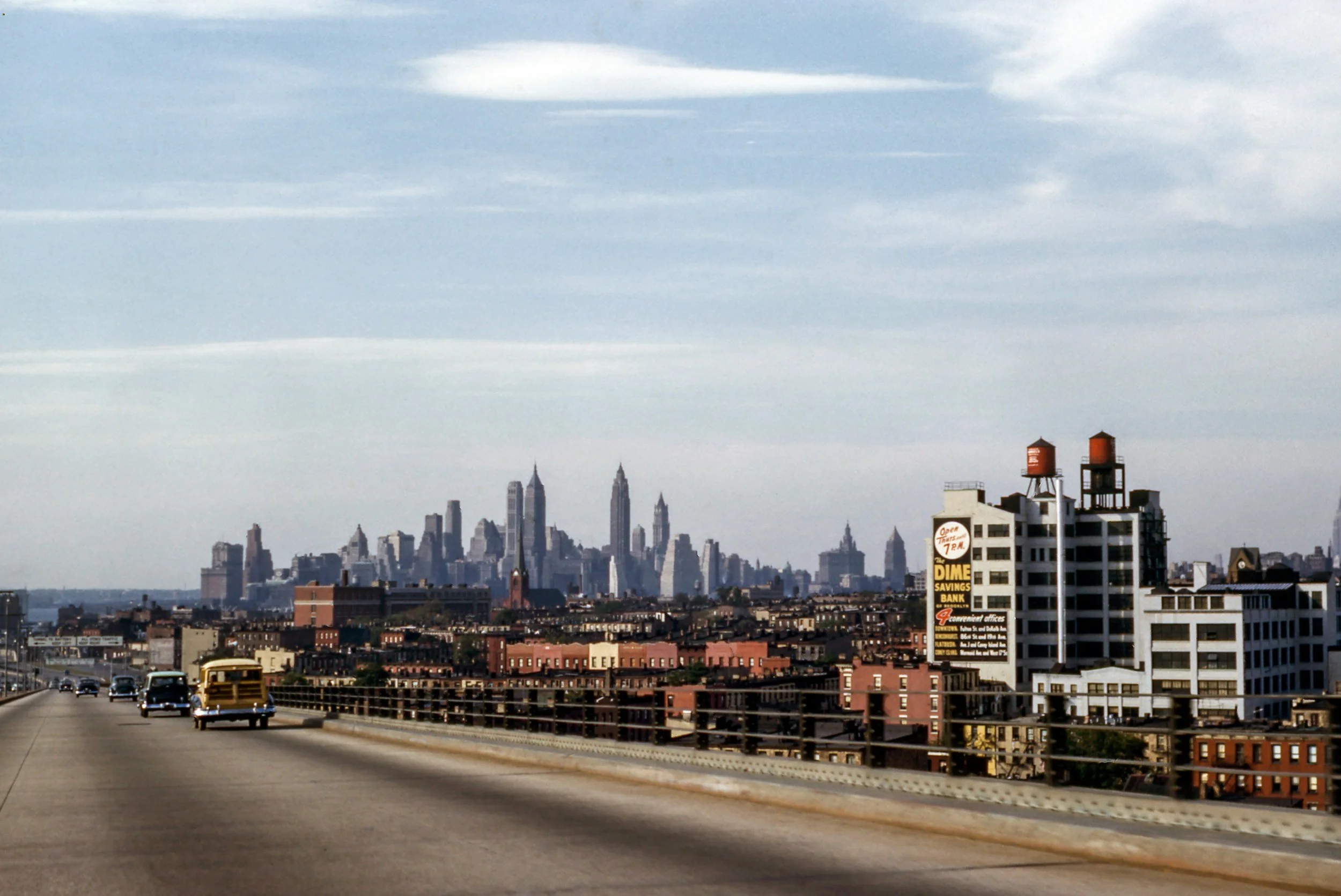 City skyline view of New York City seen from a highway with cars, including a yellow school bus, in the foreground. The skyline features tall buildings and skyscrapers under a partly cloudy sky.
