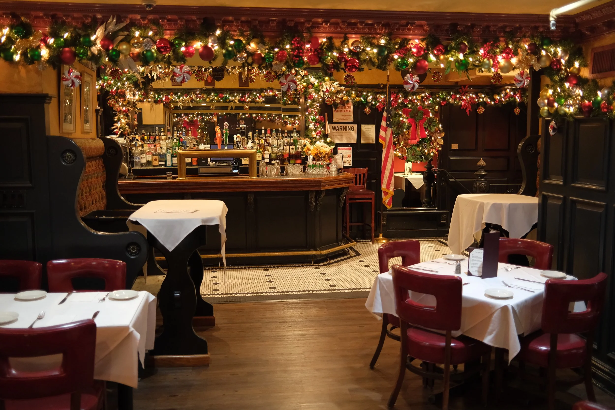 A restaurant decorated for Christmas with garlands, ornaments, and wreaths. The bar area is in the background with bottles of alcohol and festive decorations. There are tables with white tablecloths and red chairs in the foreground.
