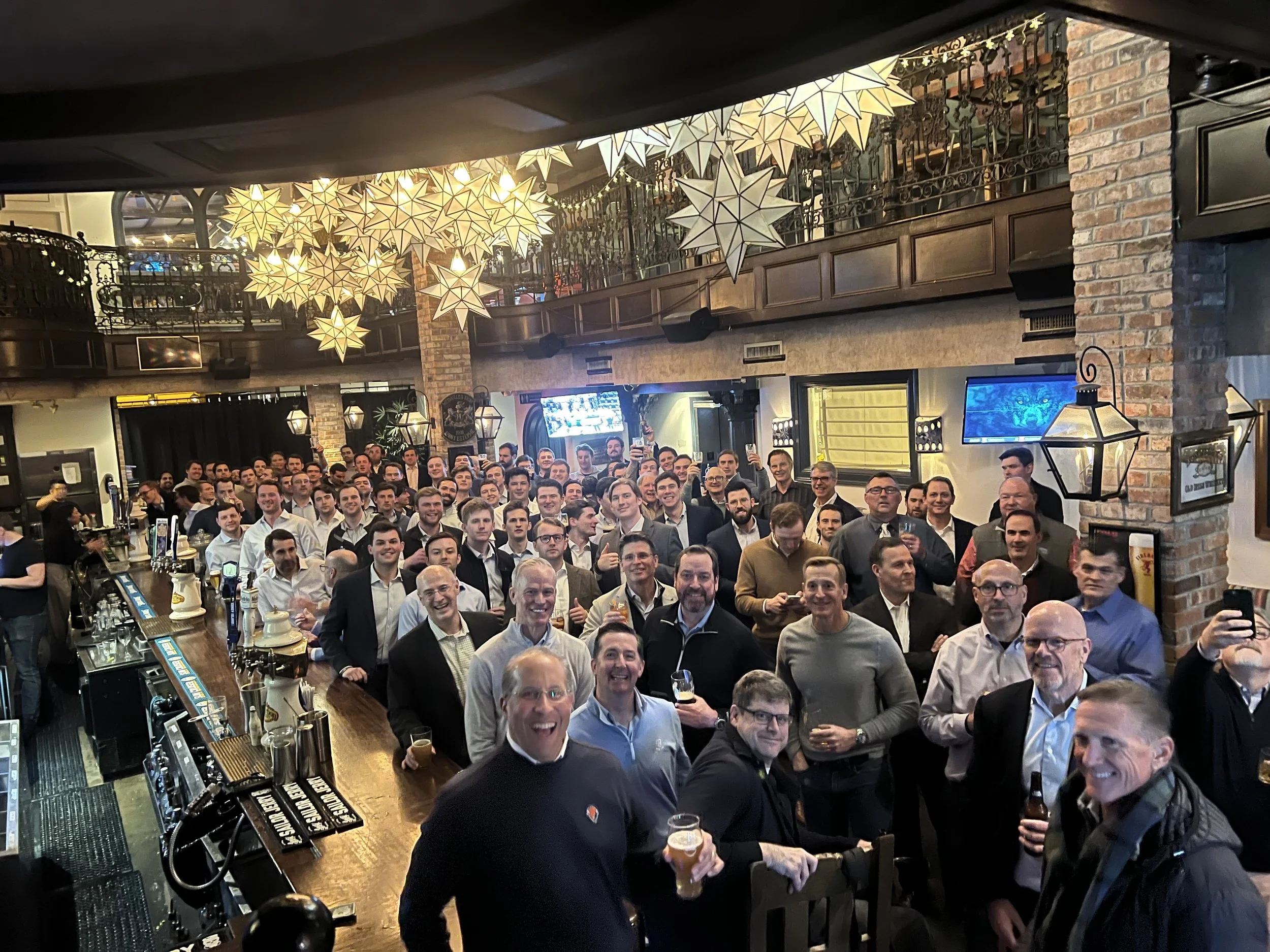 Group of people gathered inside a bar or restaurant celebrating, with some holding drinks, under decorative star-shaped party lights.