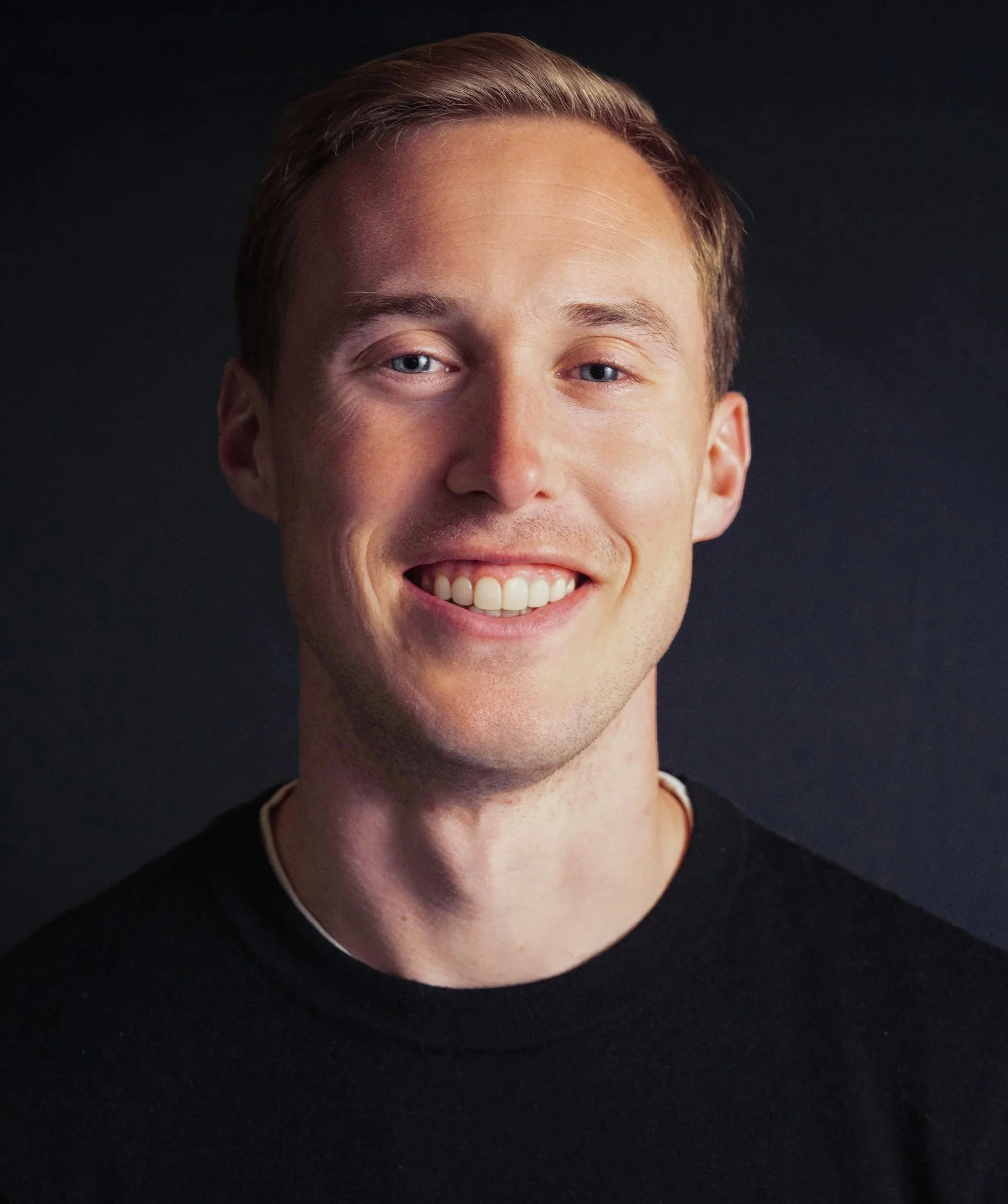Close-up portrait of a smiling man with short light brown hair, wearing a black shirt, against a dark background.