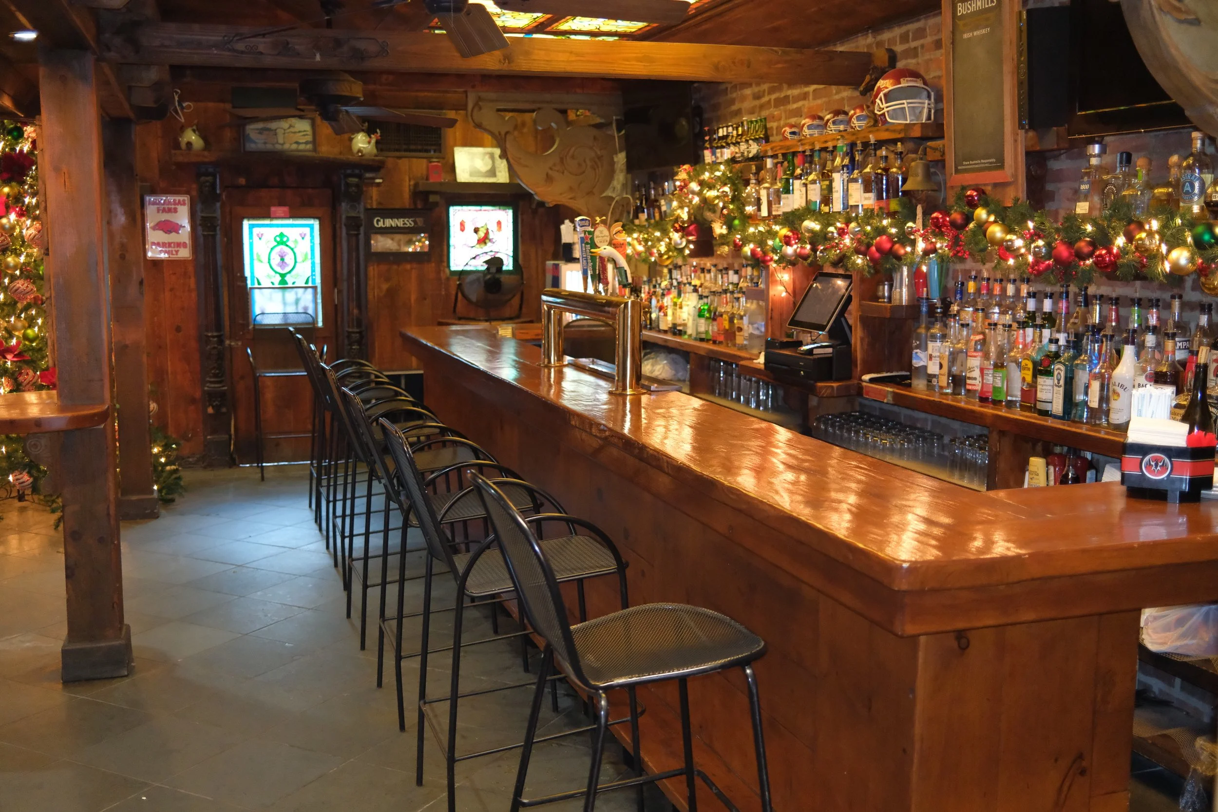 Interior of a bar decorated with Christmas ornaments and lights, with a wooden counter, barstools, shelves filled with bottles, and stained glass windows.