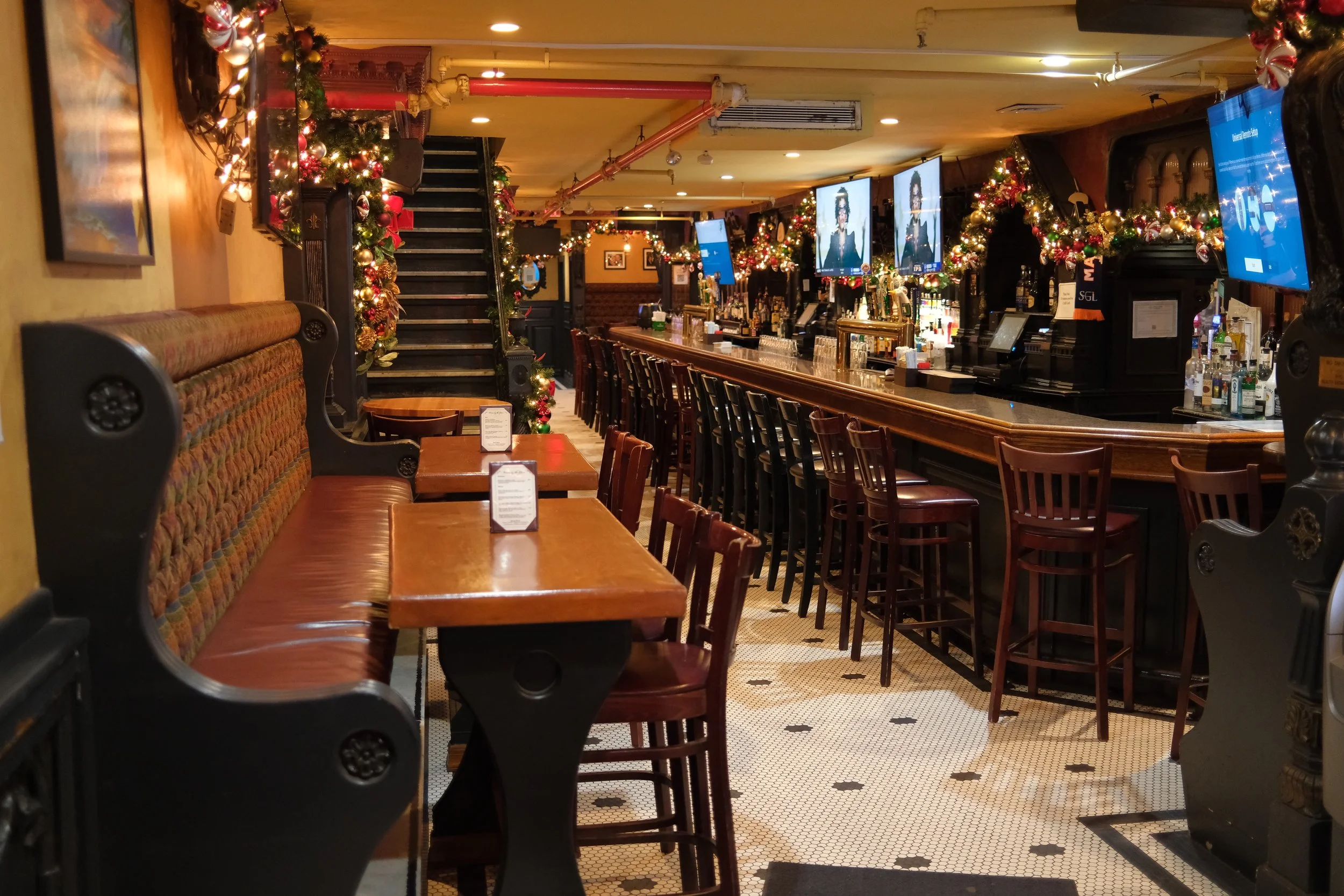 Interior of a bar decorated for Christmas with garlands, ornaments, and lights. There are tables and chairs on the left, a long bar counter on the right with stools, and multiple TV screens mounted above the bar displaying a woman in a black outfit.