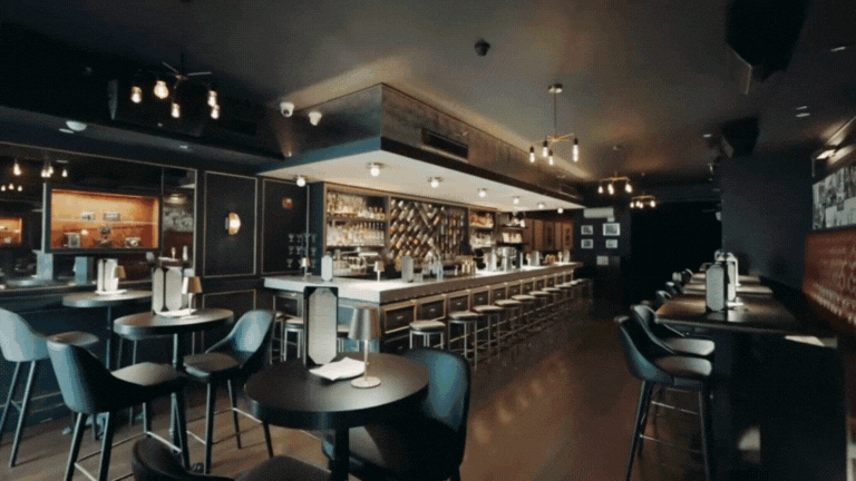 Interior of a modern bar or restaurant with high tables, bar stools, and a well-stocked bar counter.
