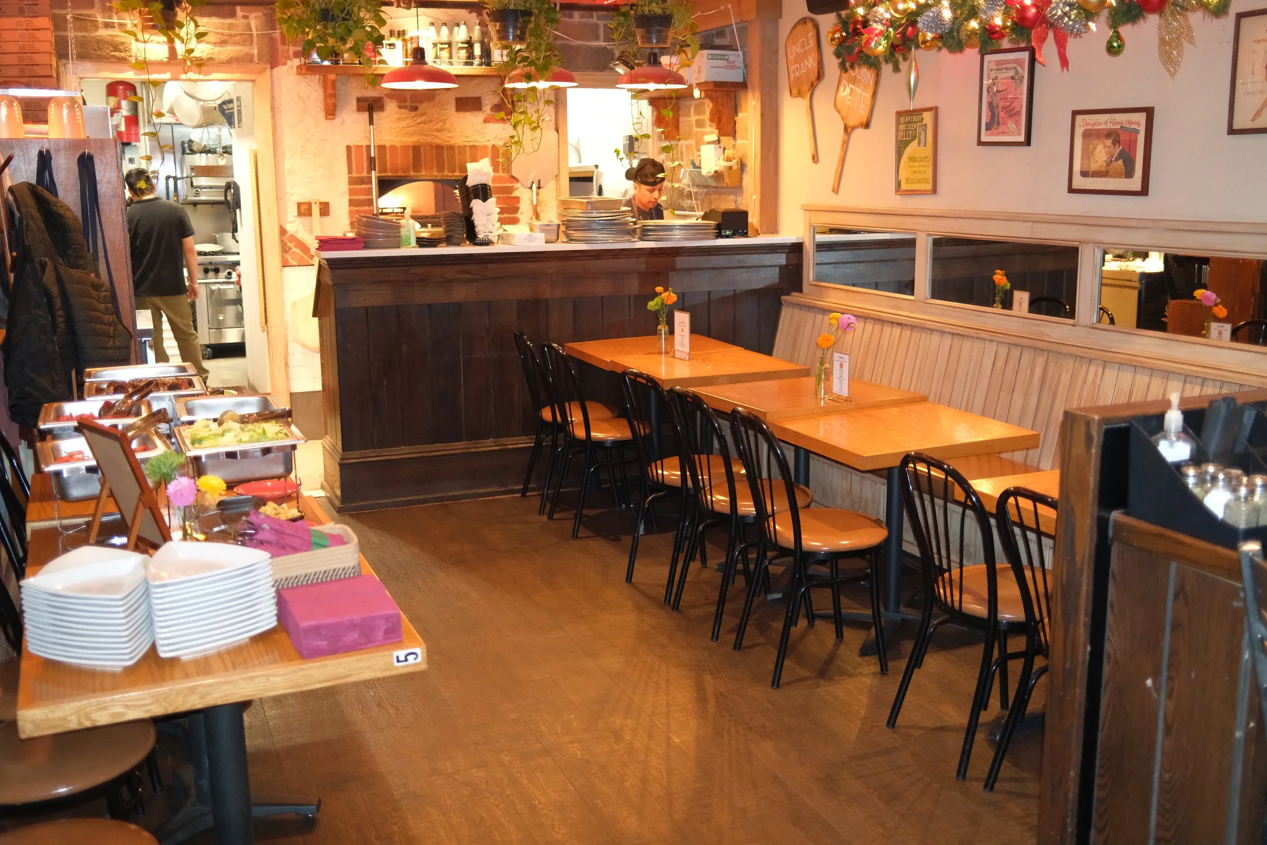Cozy restaurant interior with wooden tables and black chairs, decorated with small flower vases, and a buffet setup with dishes and plates on a side table. The open kitchen area shows staff working, and the wall is decorated with framed posters and h