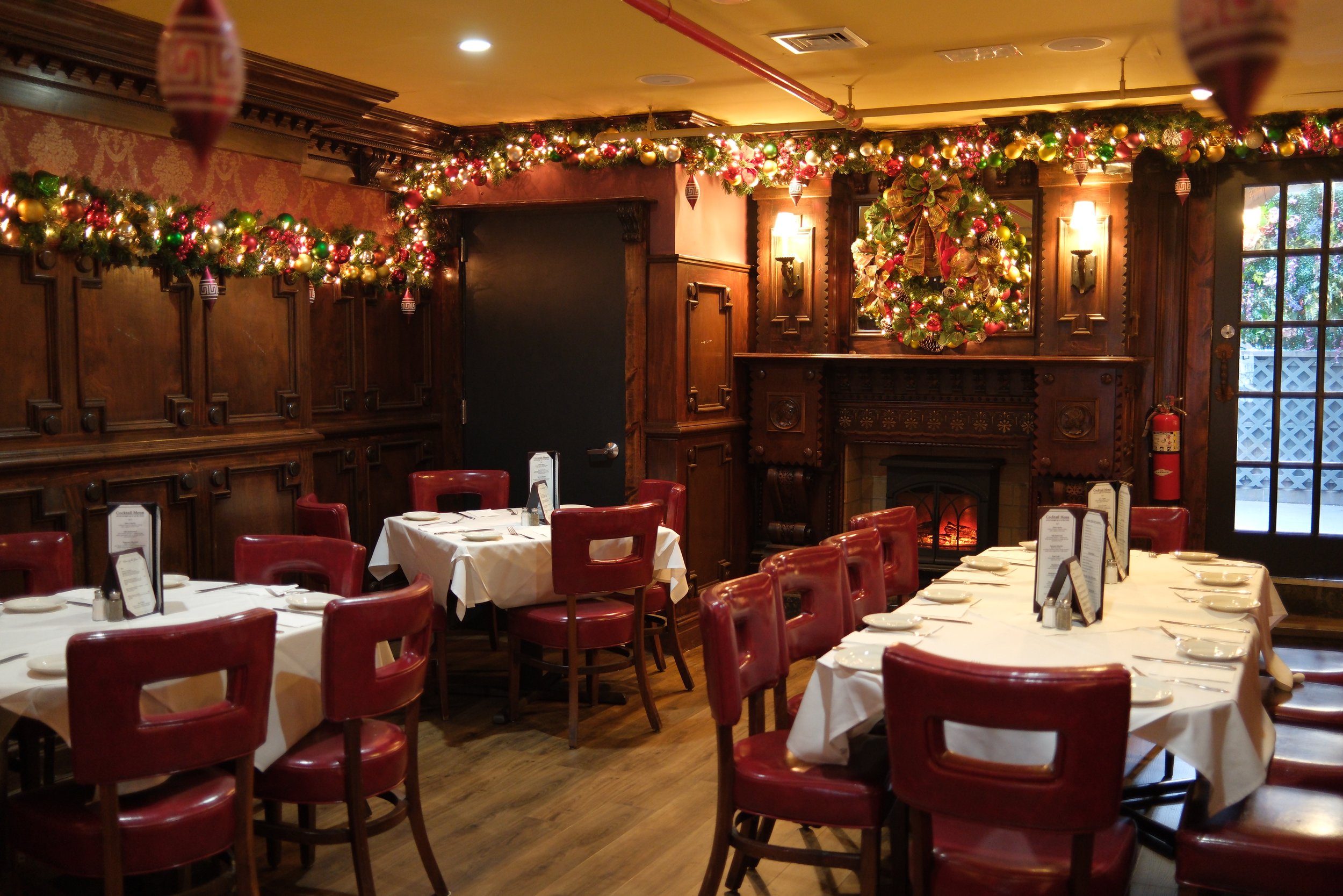 Christmas decorated restaurant with tables set for dining, adorned with garlands, ornaments, and a wreath above the fireplace, featuring wood paneling and a fire.