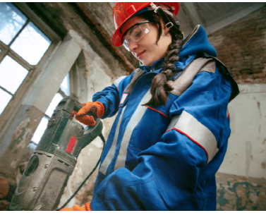 Female construction worker wearing a red safety helmet, protective glasses, and a blue jacket using a power drill inside a building under construction.