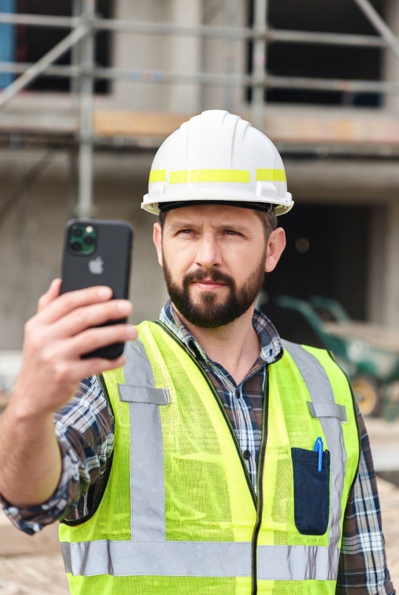 A man wearing a yellow safety vest and a white construction helmet takes a selfie at a construction site.