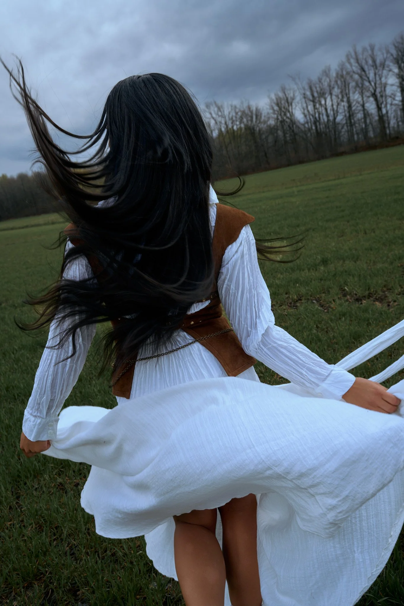 Femme en robe blanche assise dans un champ vert, cheveux noirs longs soufflés par le vent, ciel nuageux en arrière-plan. Photo prise par Maison Toki pour Maison Préfontaine. pour la collection printemps été 2026.