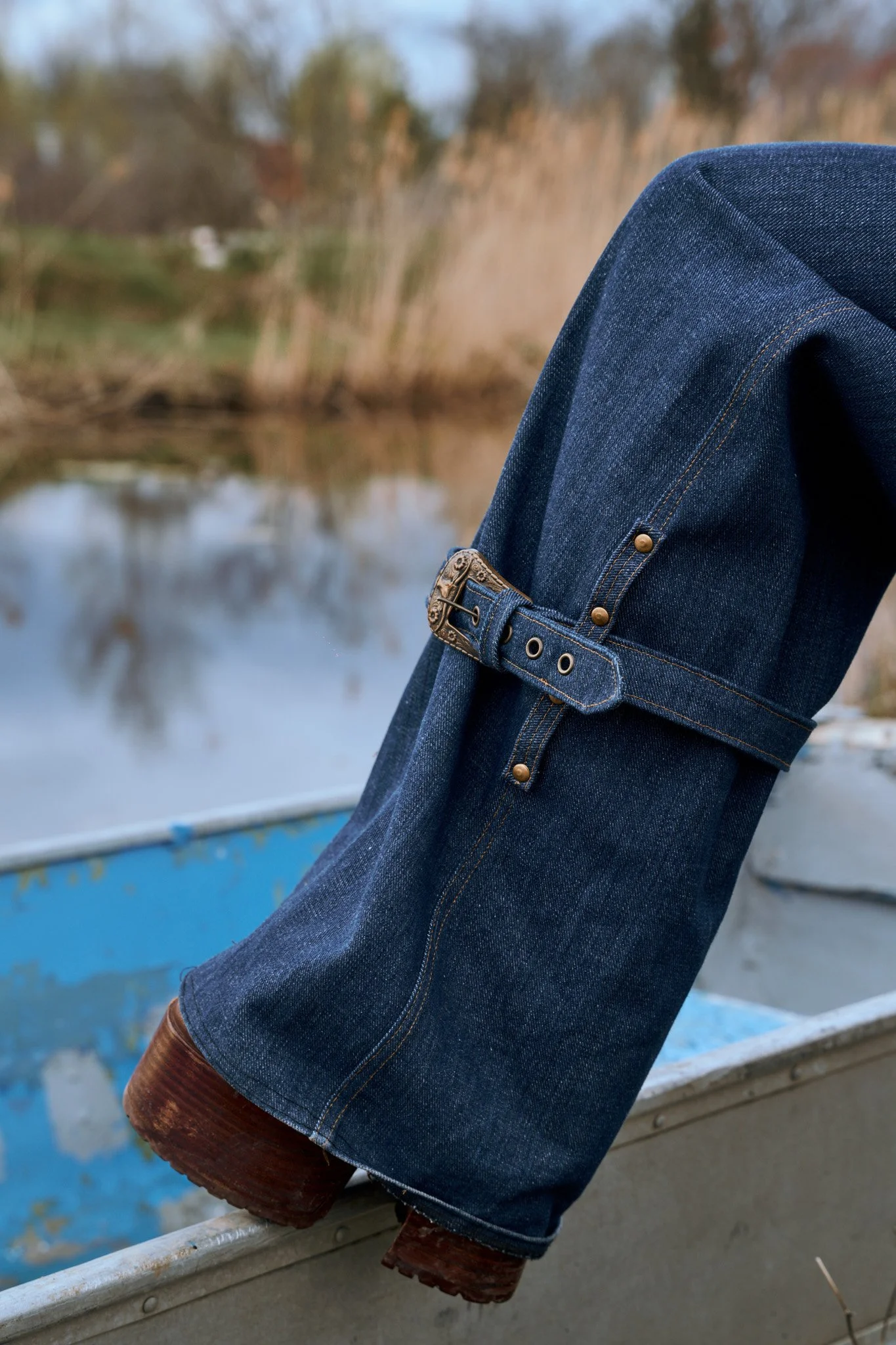 Une personne portant un pantalon en jean foncé avec des bottes en cuir marron, assise ou appuyée contre une barrière en métal près d'une étendue d'eau, avec des arbres en arrière-plan. . Photo prise par Maison Toki pour Maison Préfontaine.