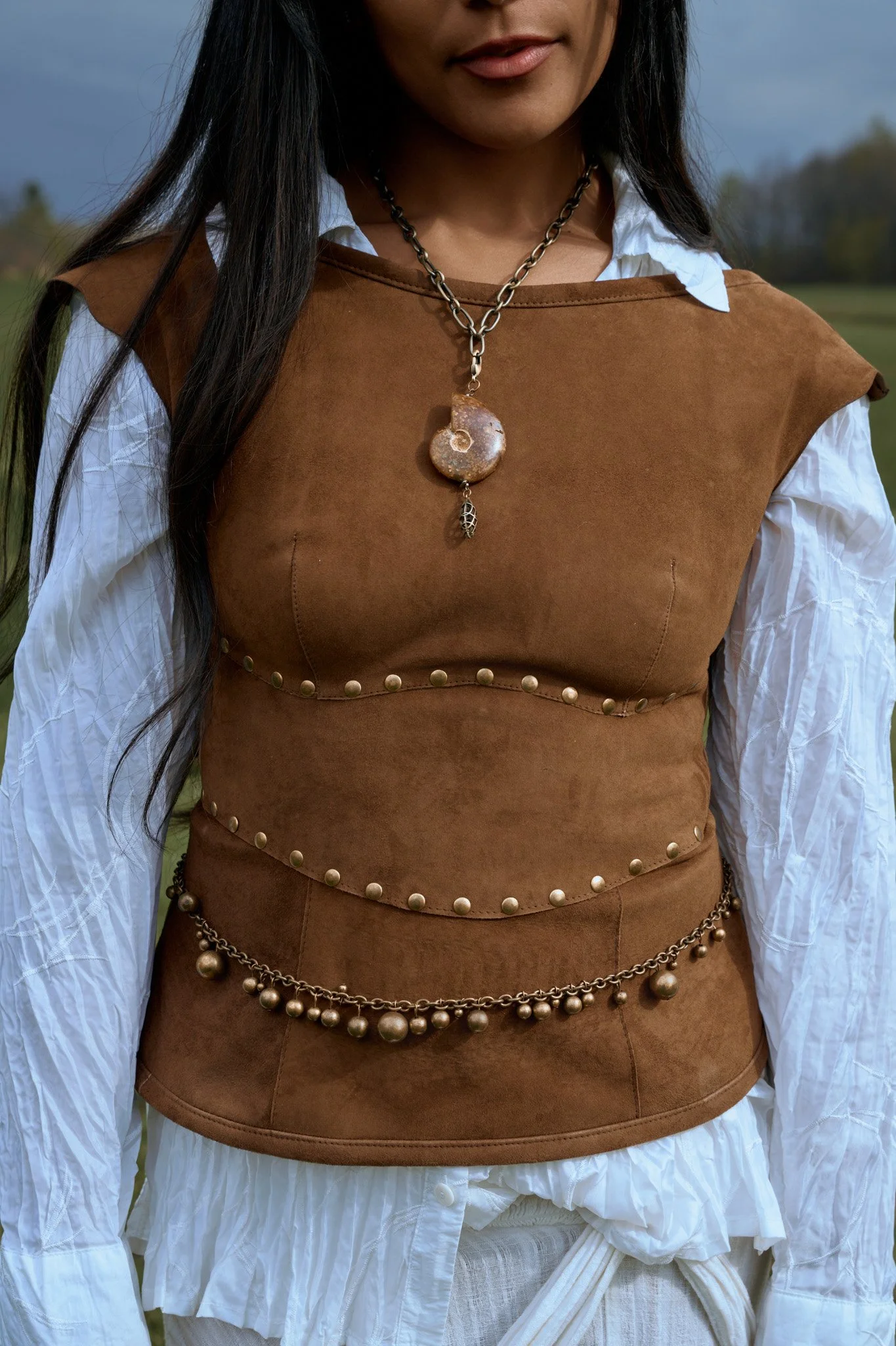 Une femme portant un haut en cuir marron avec des détails de perles dorées, un collier en chaîne métallique avec un pendentif en forme de coquillage et un autre en forme de feuille. Photo prise par Maison Toki pour Maison Préfontaine.
