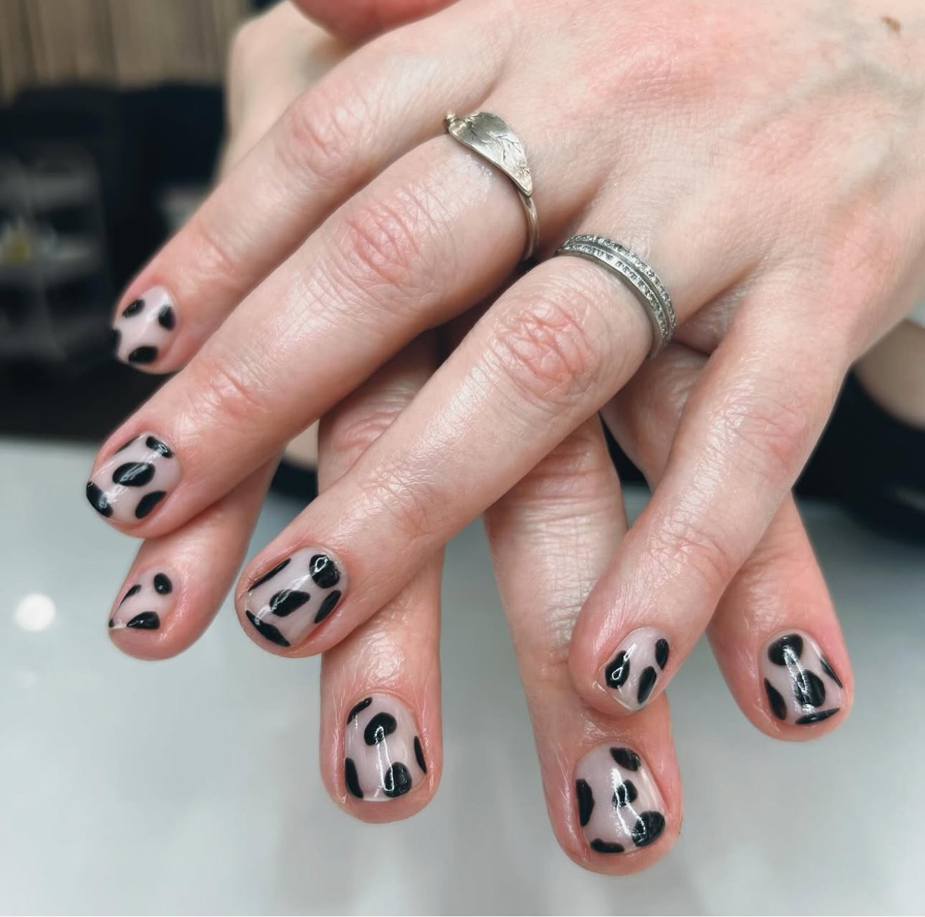 Close-up of a person's hands with black and beige leopard print nail polish, wearing two rings—one plain silver and the other with stones.
