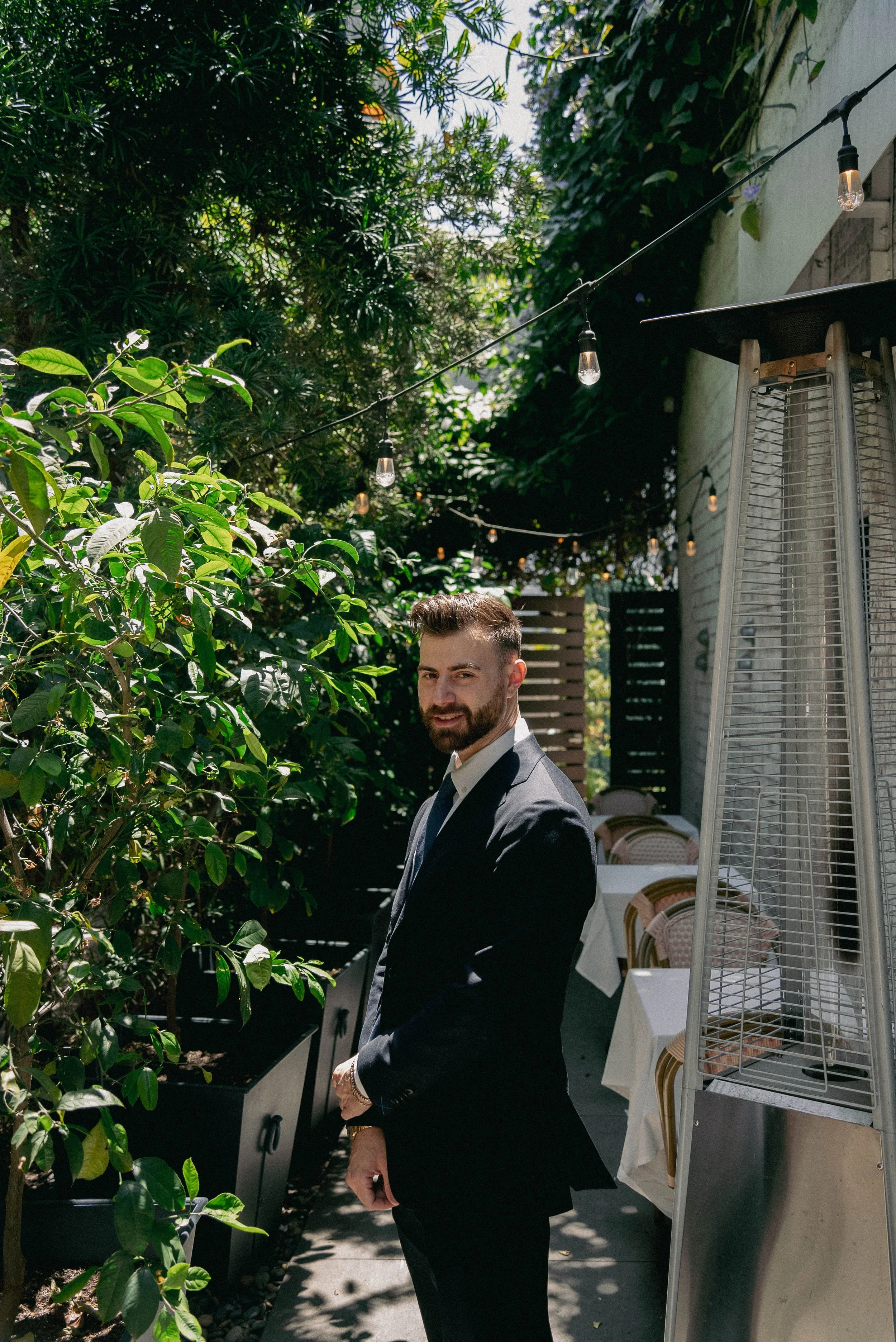 A man in a dark suit with a white shirt standing outdoors near green plants and trees, with string lights above and a patio heater to the right, in a garden or backyard setting.