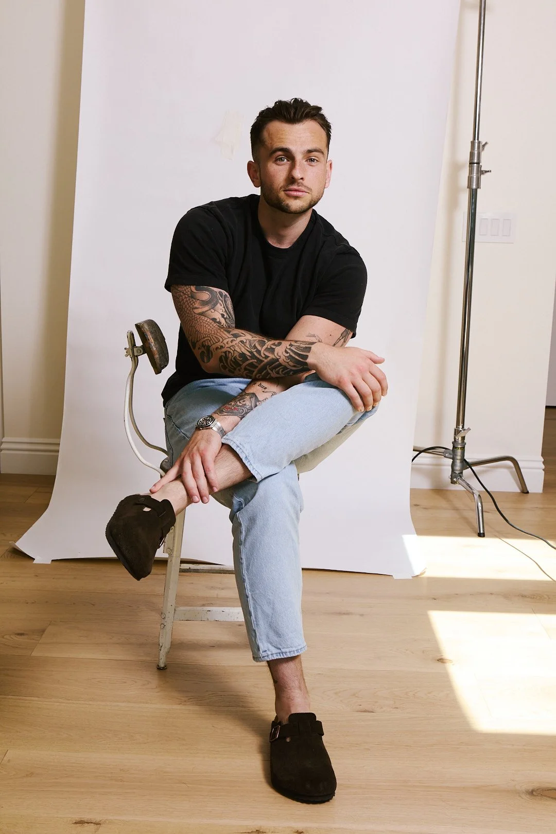 A man with tattoos, wearing a black t-shirt, light blue jeans, and black slippers, sits on a chair in front of a white backdrop in a photography studio.