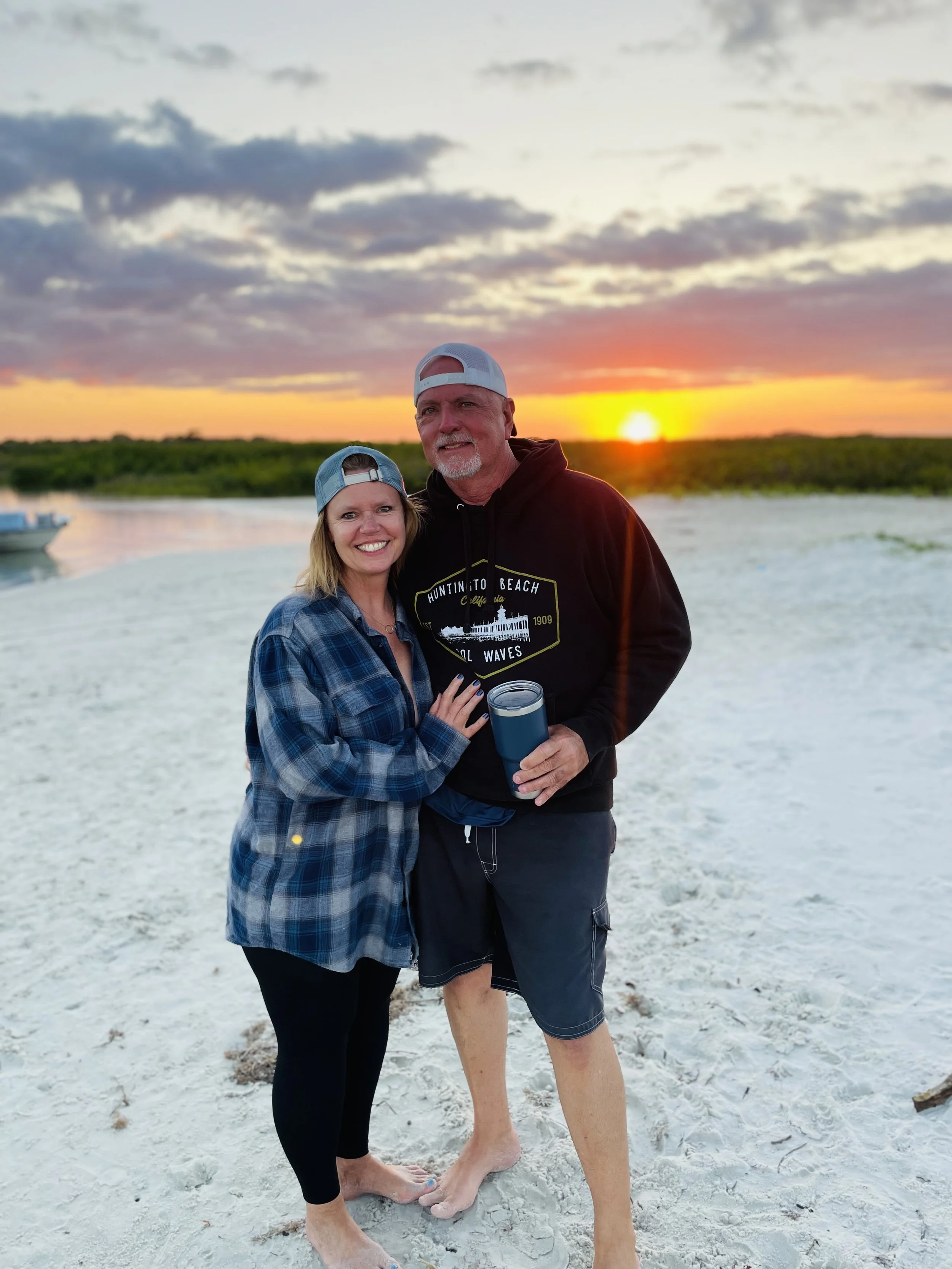 A couple standing on a beach during sunset, both wearing casual beachwear including caps and holding a beverage, smiling at the camera. The sky shows a colorful sunset with some clouds.