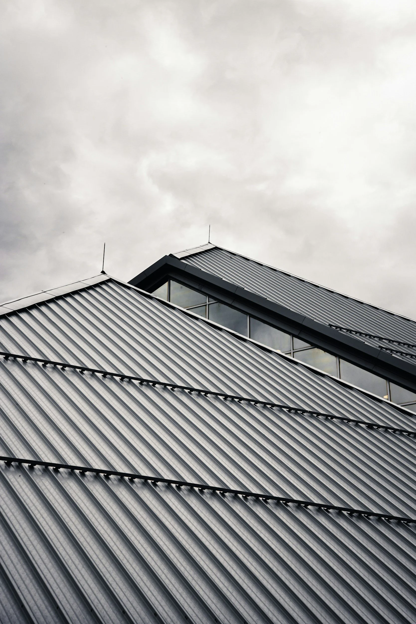 Close-up of a modern building with an angular, metallic roof and large glass windows, under an overcast sky.