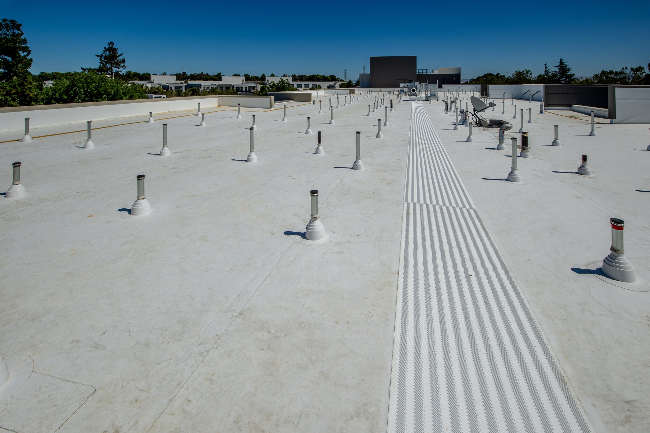 Rooftop with white surface, multiple vent pipes, a satellite dish, and a tactile walking surface indicator strip, with trees and buildings in the background under a clear blue sky.