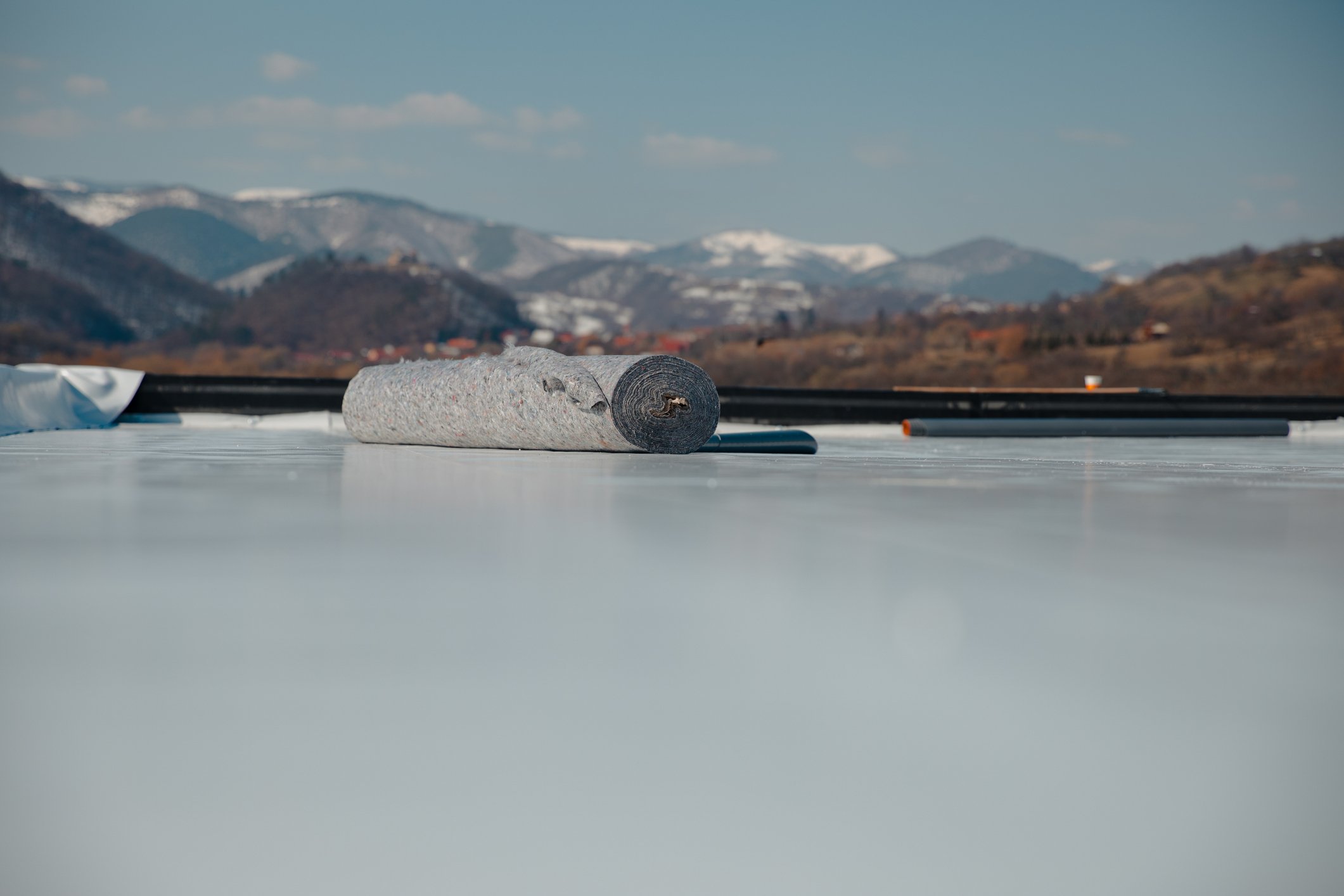 A close-up of a roll of roofing felt or underlayment lying on a flat surface, with mountains and cloudy sky in the background.