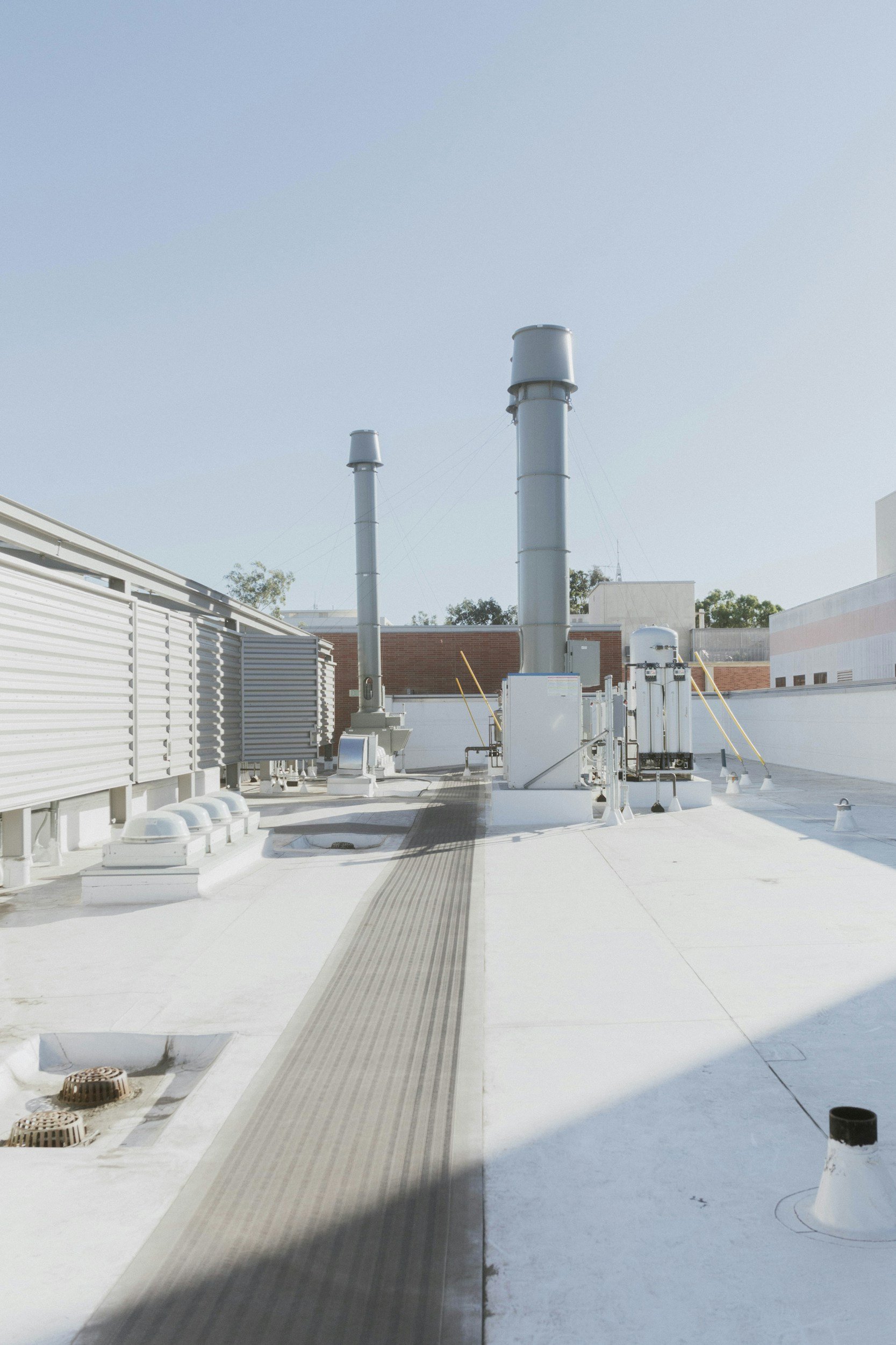 Rooftop with multiple vent pipes, HVAC units, and a walkway on a building with a white surface and a clear sky.