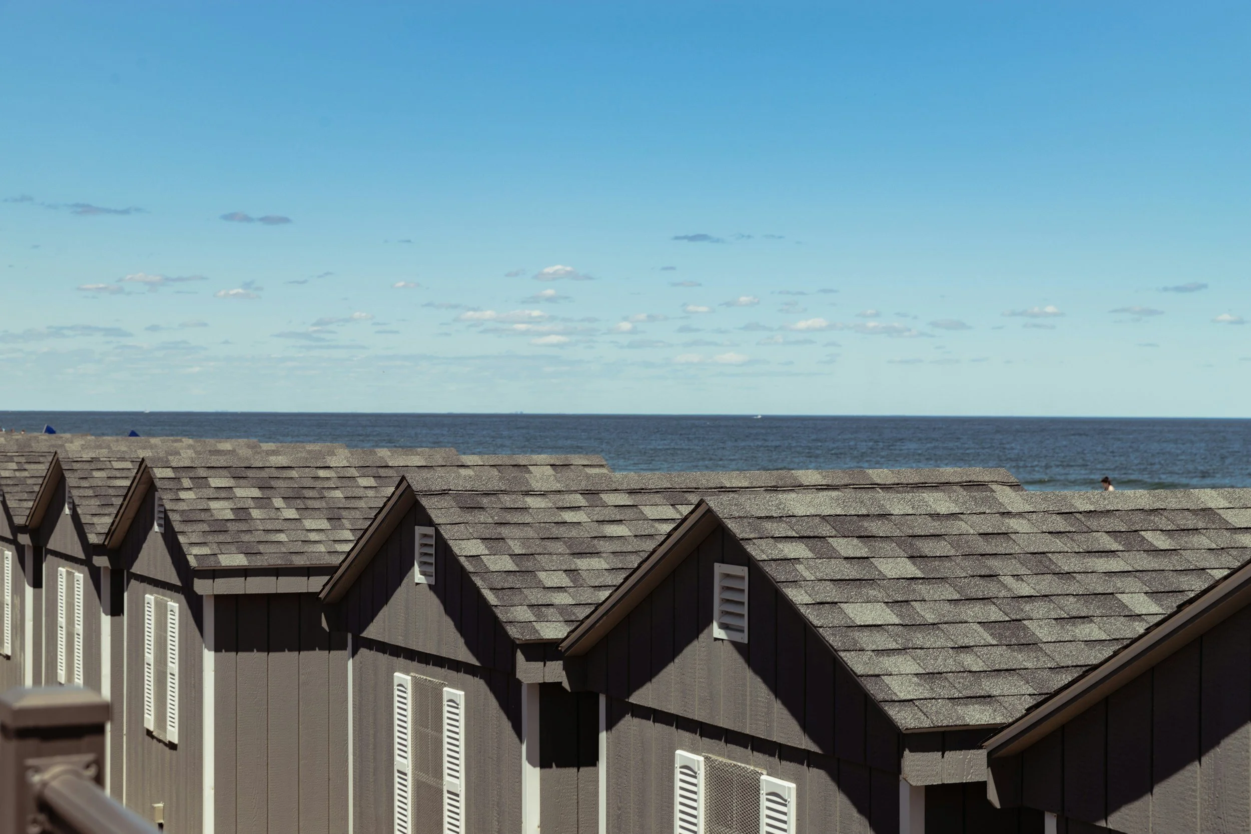 A row of seaside houses with gray shingle roofs in the foreground, with the ocean and sky in the background, and a person walking on the beach.
