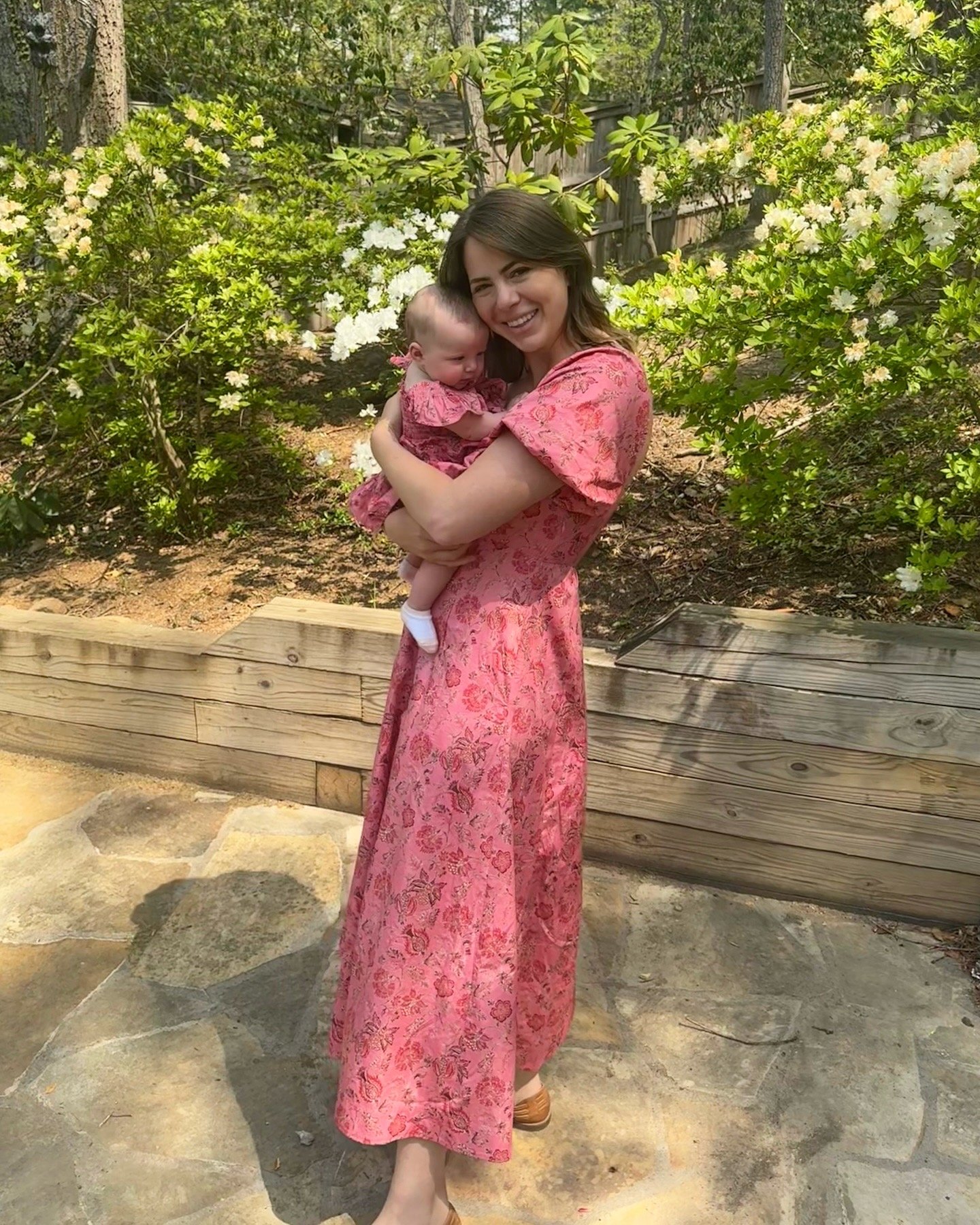 A mother holding her baby, both wearing matching pink floral dresses, stands outdoors on a sunny day with green foliage and white flowers in the background.