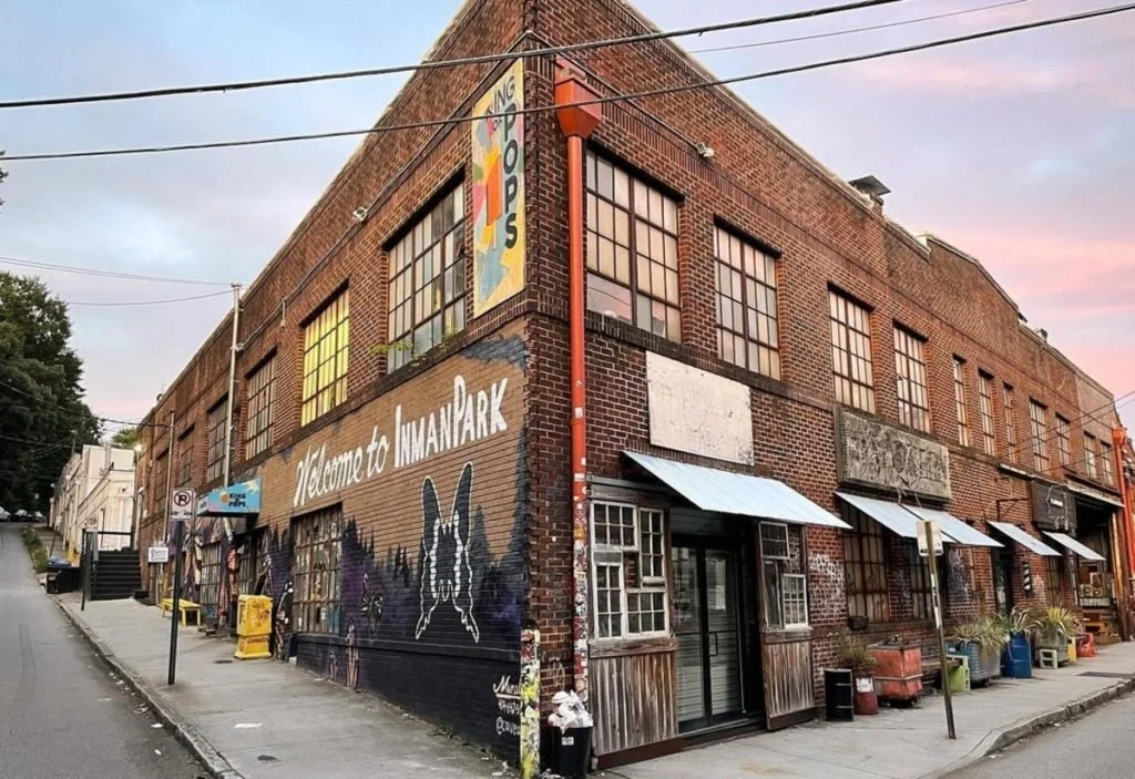 A brick building on a street corner with Welcome to Inman Park mural and colorful shop signs, awnings, and outdoor seating.