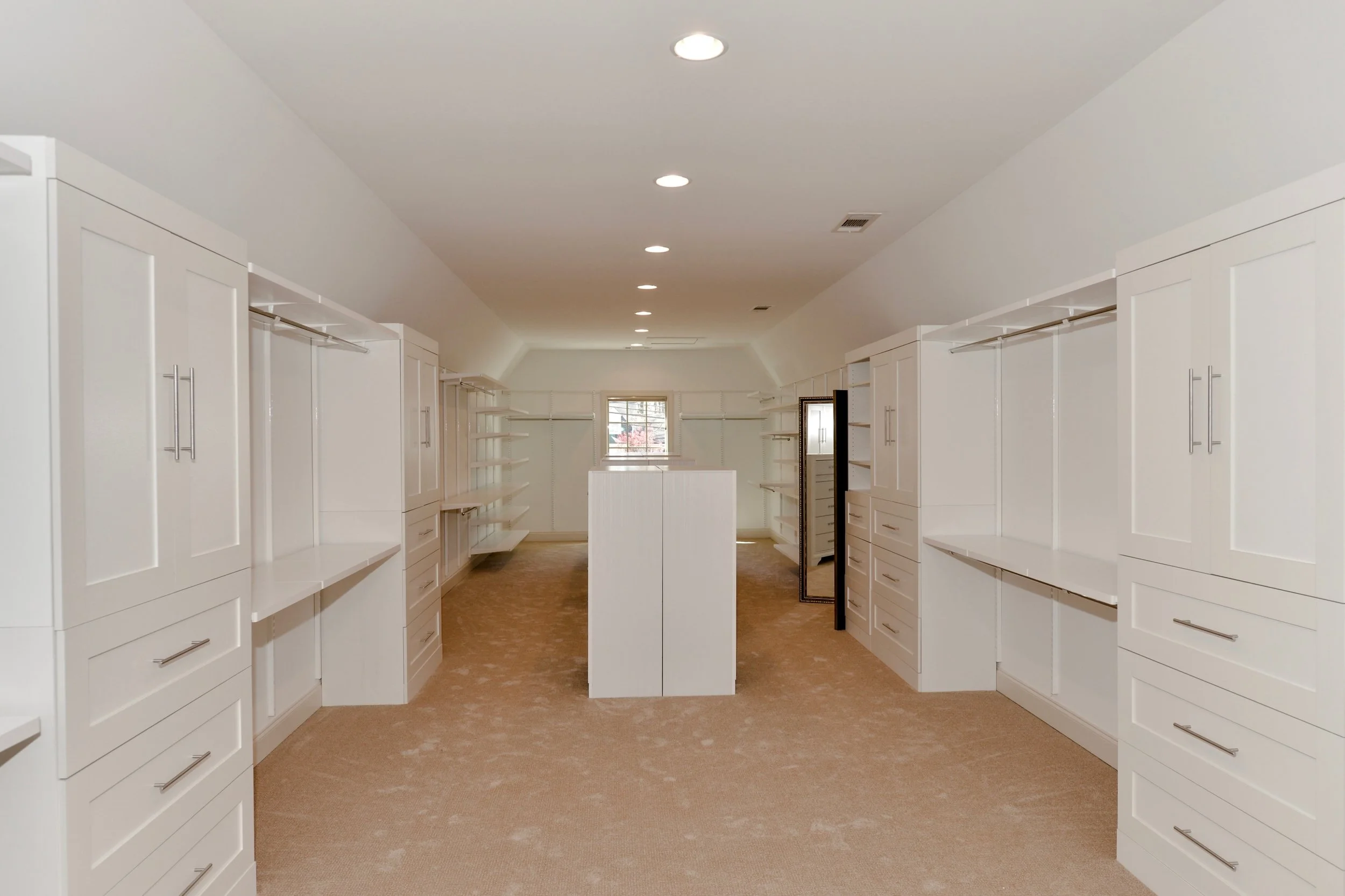 Empty walk-in closet with white cabinetry and open shelves, beige carpet, ceiling lights, and a small window at the end
