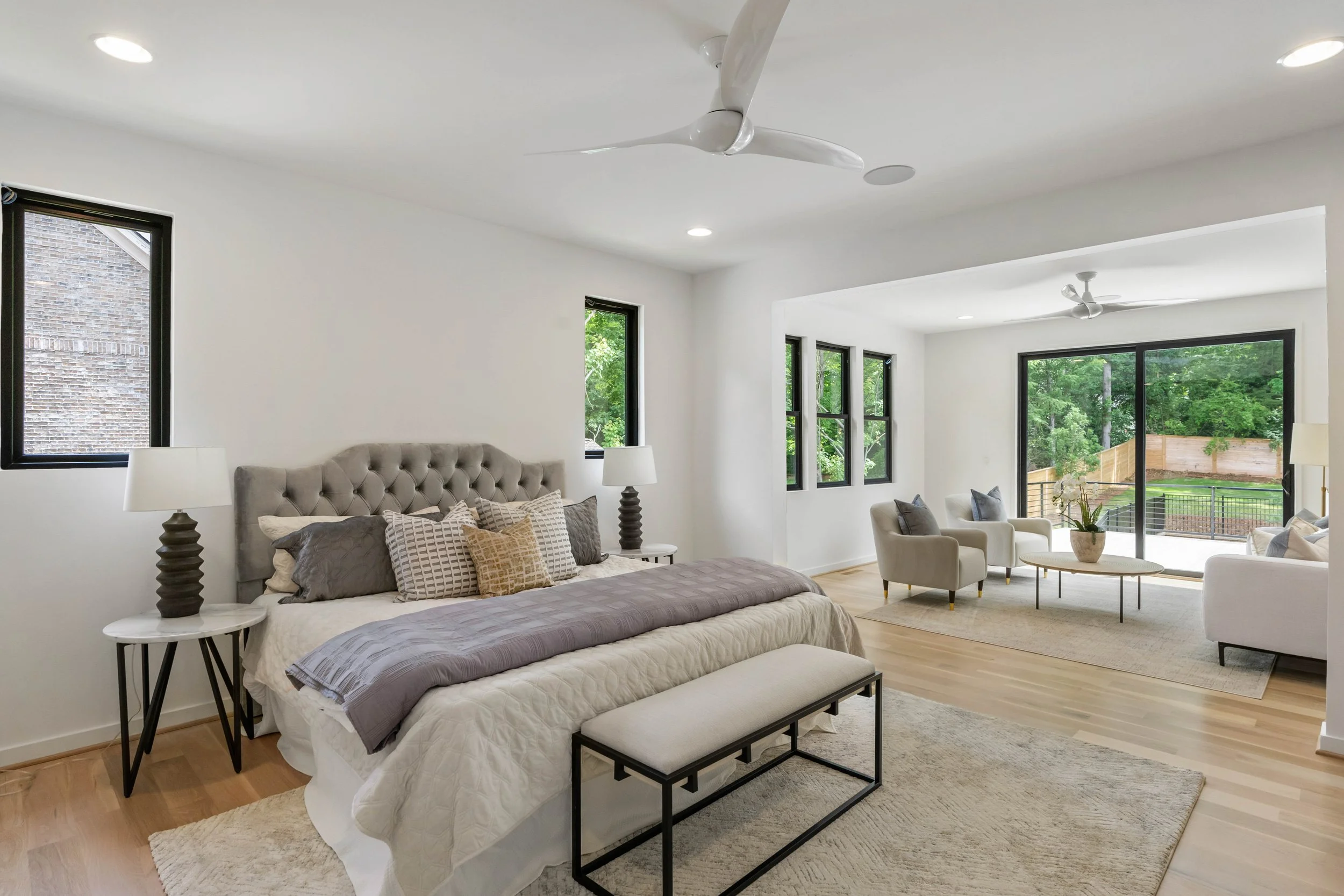 Modern bedroom with neutral color palette, tufted gray headboard, multiple patterned pillows, two bedside lamps, and a bench at the foot of the bed. Adjacent living area features white armchairs, a coffee table, large sliding glass door, and green ou