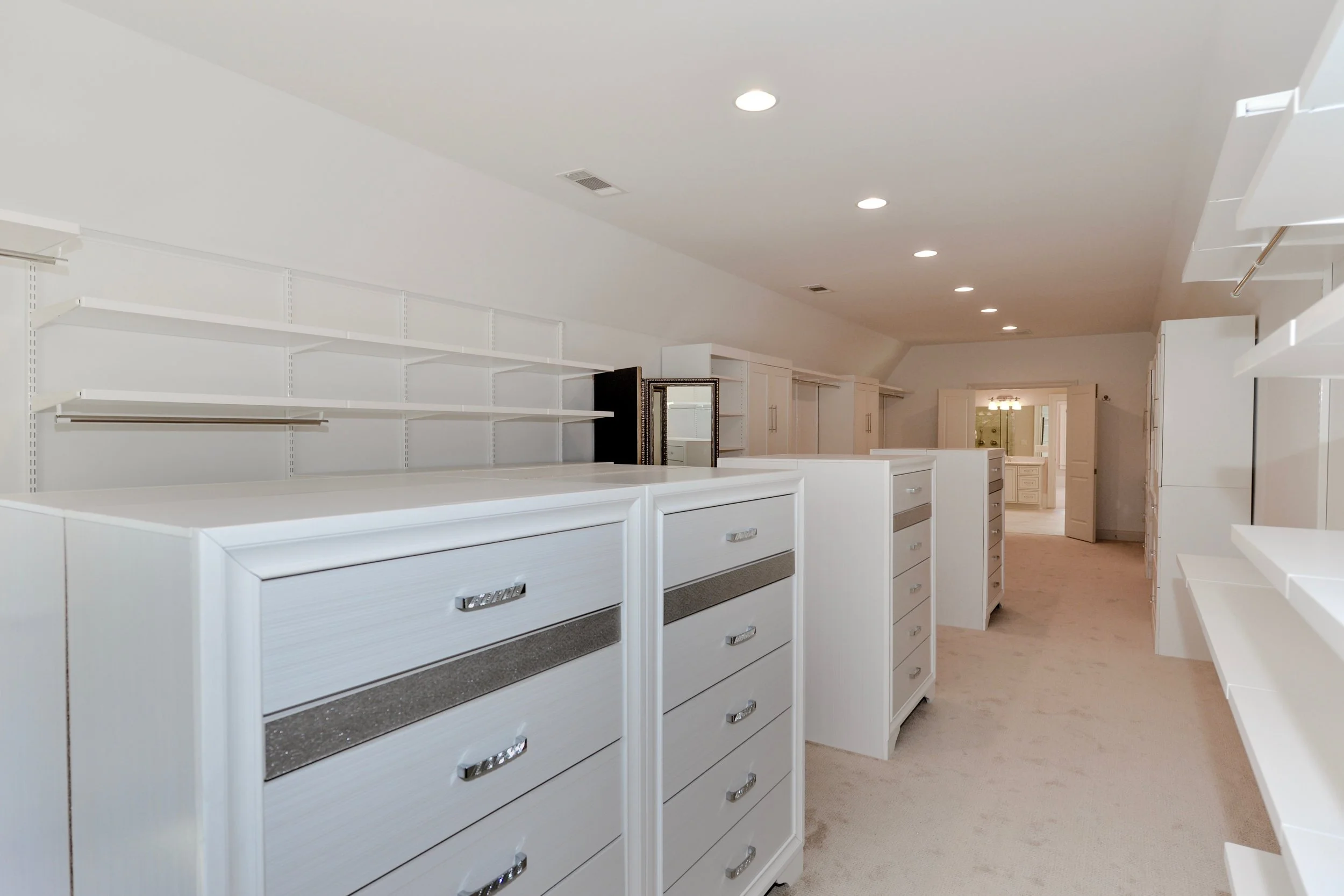 Empty walk-in closet with white built-in cabinets and open shelving, carpeted floor, located in an attic space with sloped ceiling and recessed lighting.
