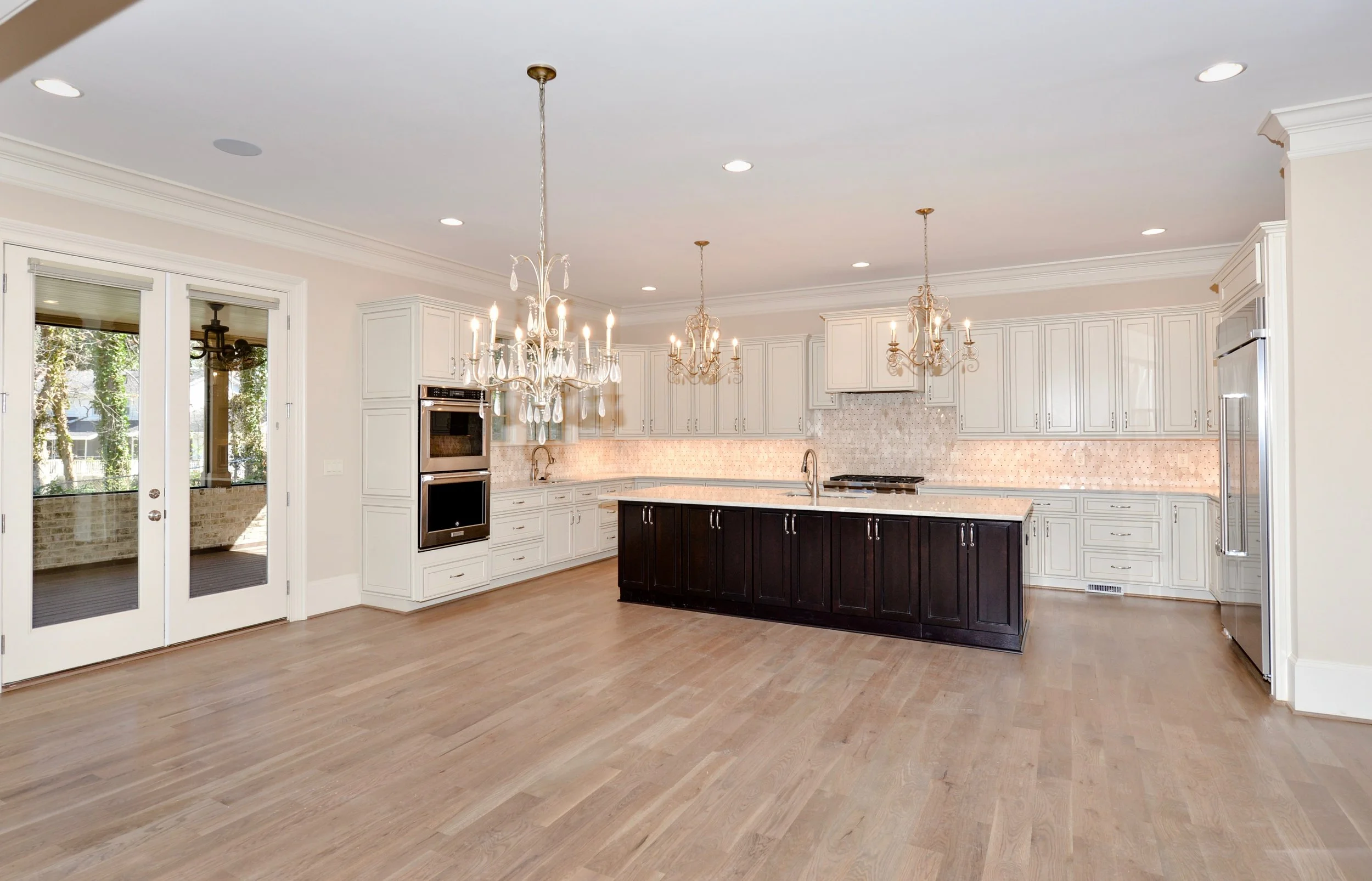 Spacious kitchen with white cabinets, a dark central island, chandeliers, stainless steel appliances, hardwood floors, and French doors leading outside.