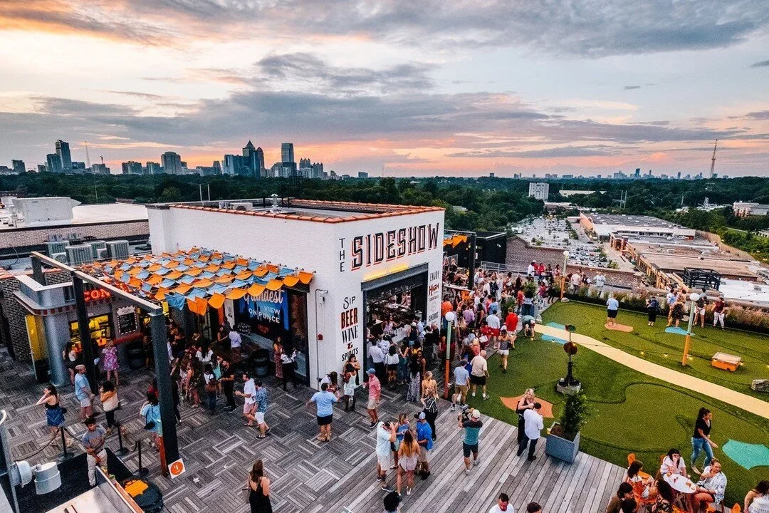 Rooftop bar at sunset with crowds, outdoor seating, lawn games, and city skyline in the background.