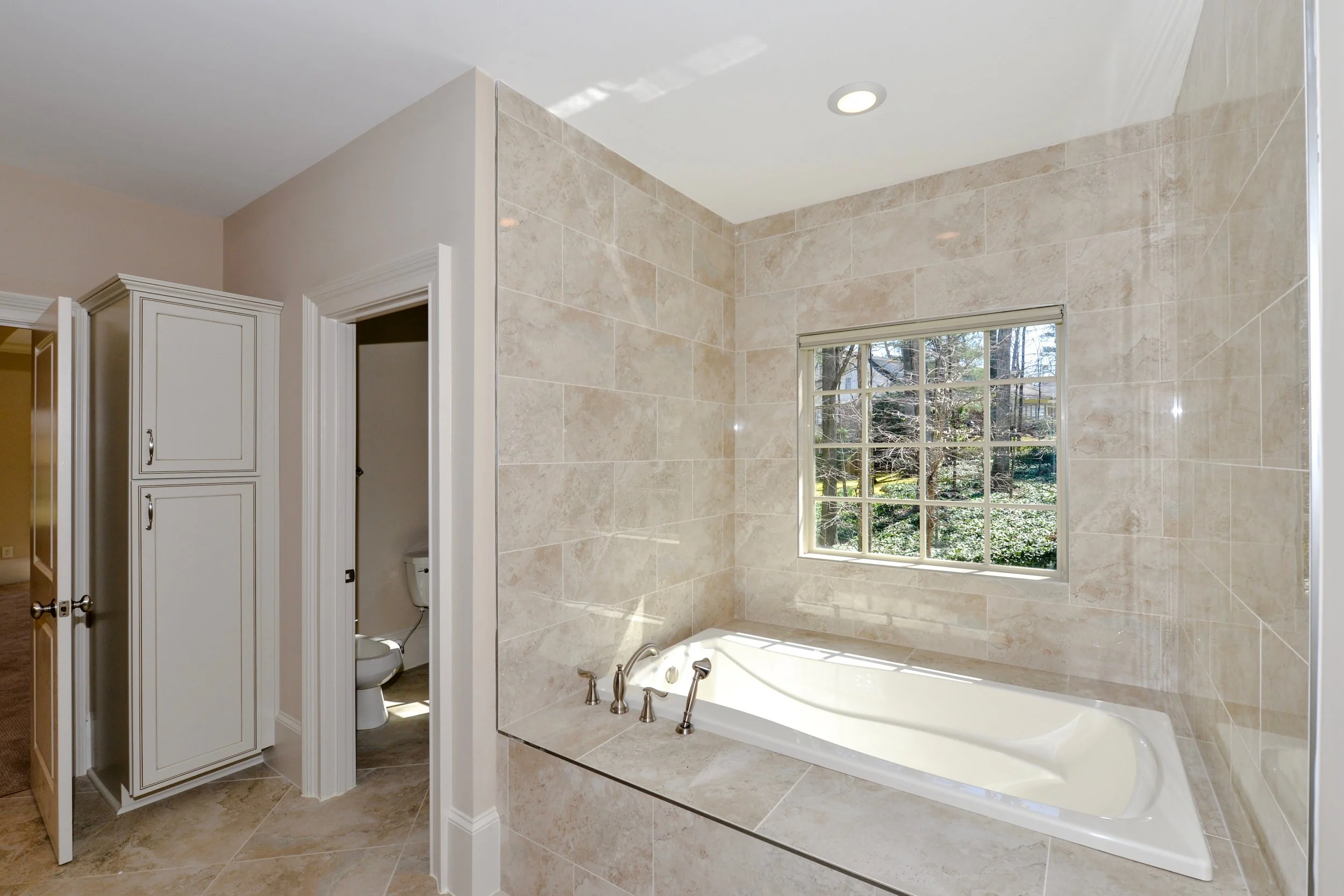 A bathroom with beige tiled walls and a large bathtub beneath a window, with a separate small toilet room visible in the background.