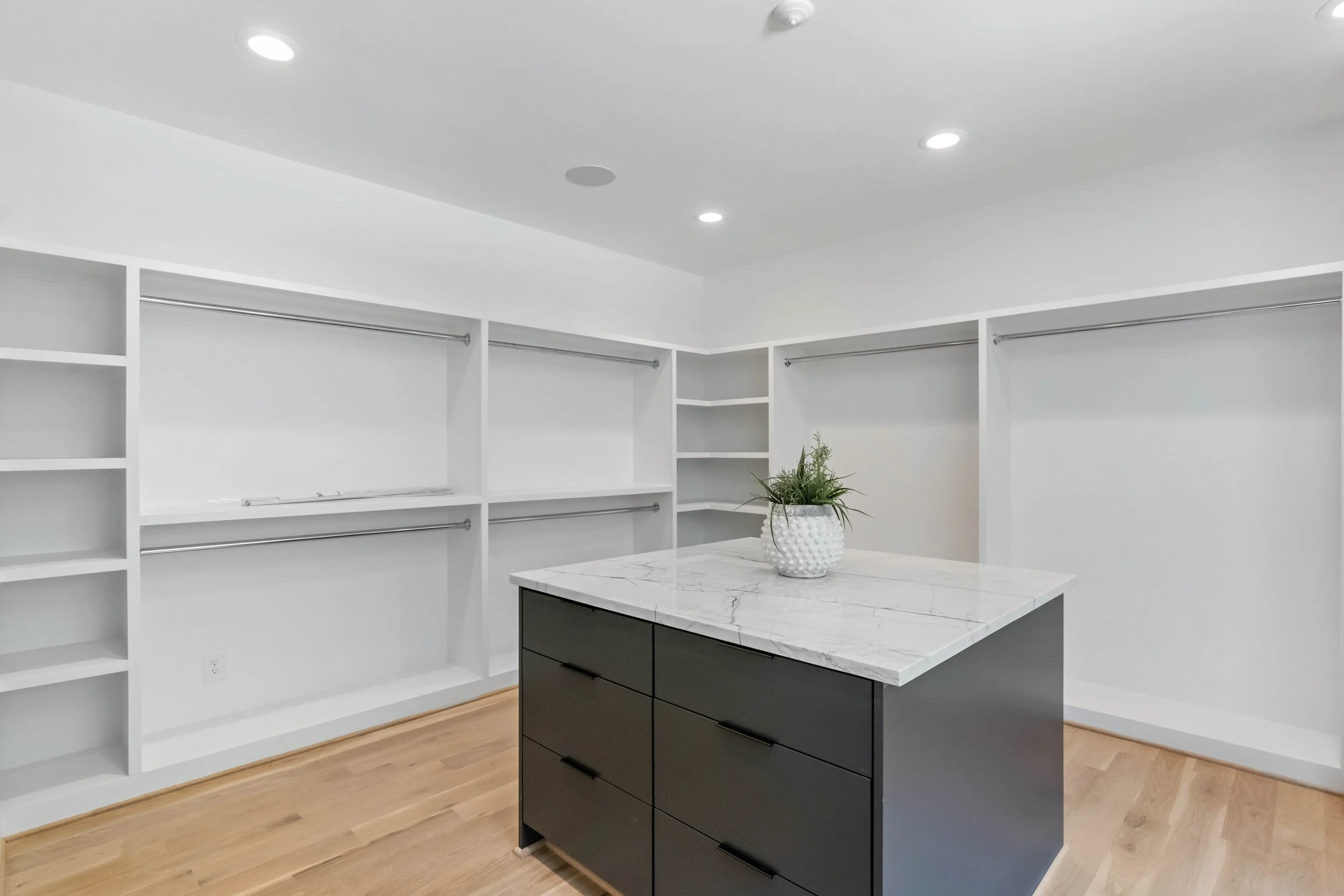 Empty walk-in closet with white built-in shelves and hanging rods, black island with white marble top, and wooden floor.