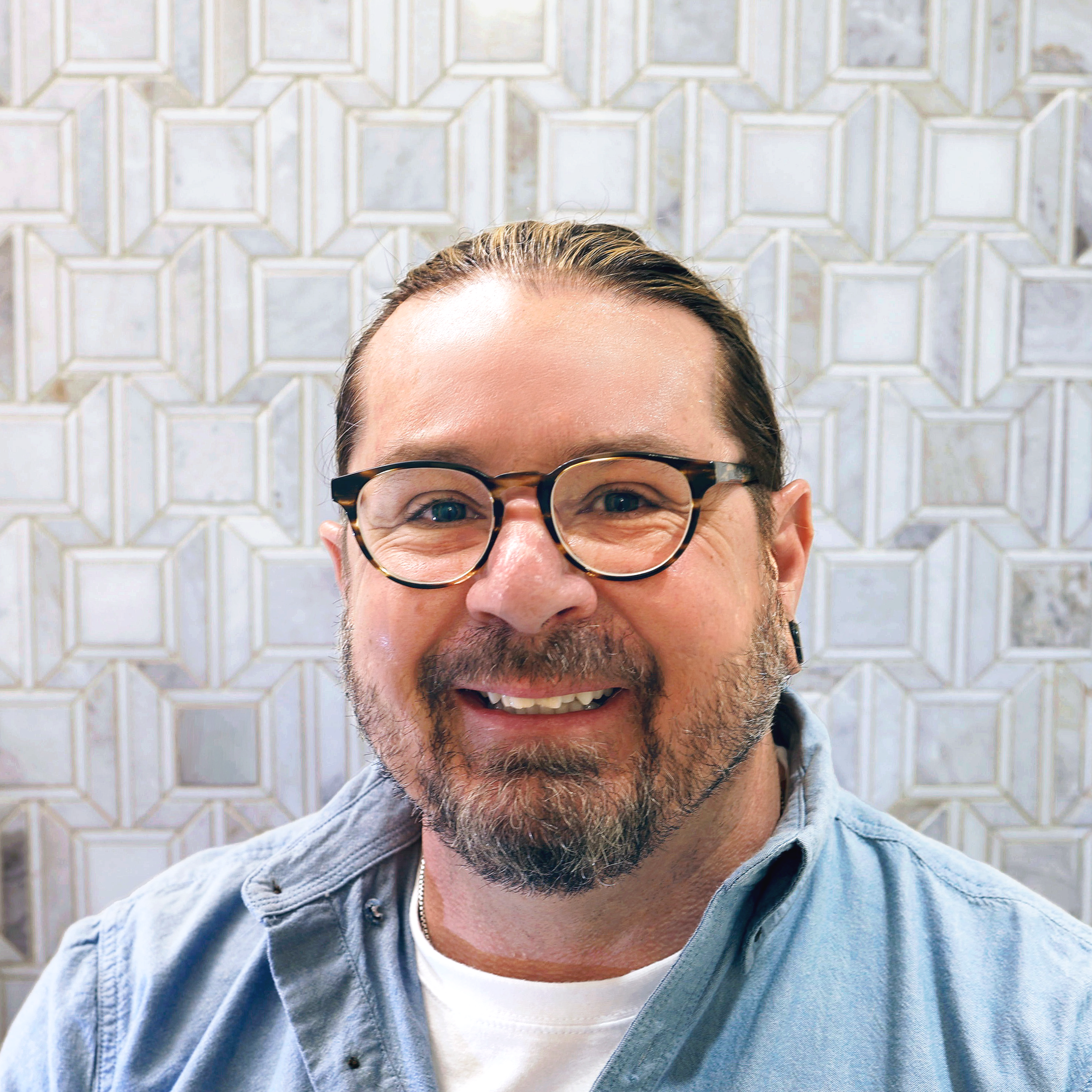 Smiling man with dark hair slicked back, beard, and earring, wearing a black shirt, standing against a light-colored tiled wall.