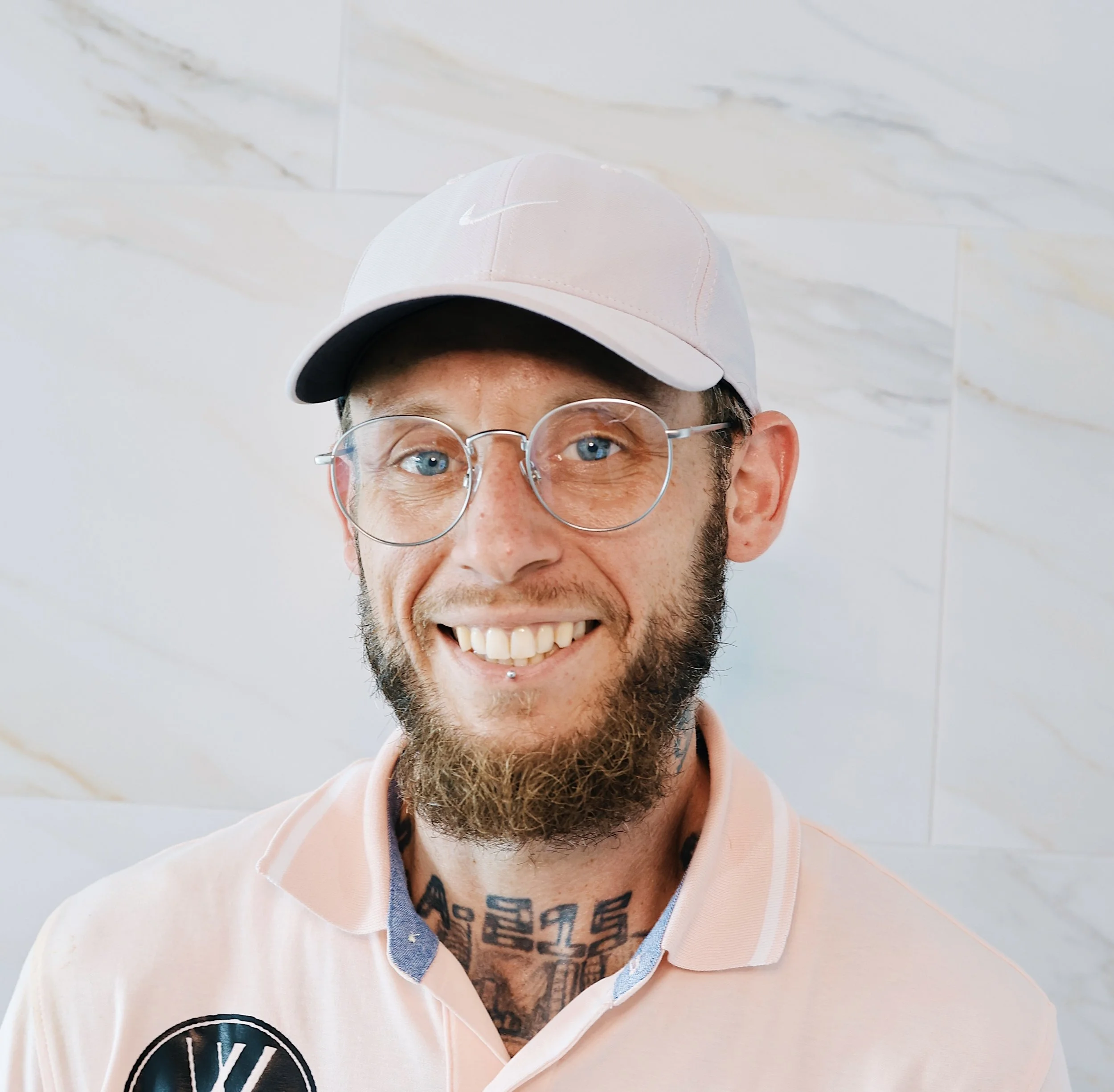 A smiling man with glasses, a beard, and tattoos wearing a pink Nike cap and a light pink polo shirt standing against a light-colored tiled wall.