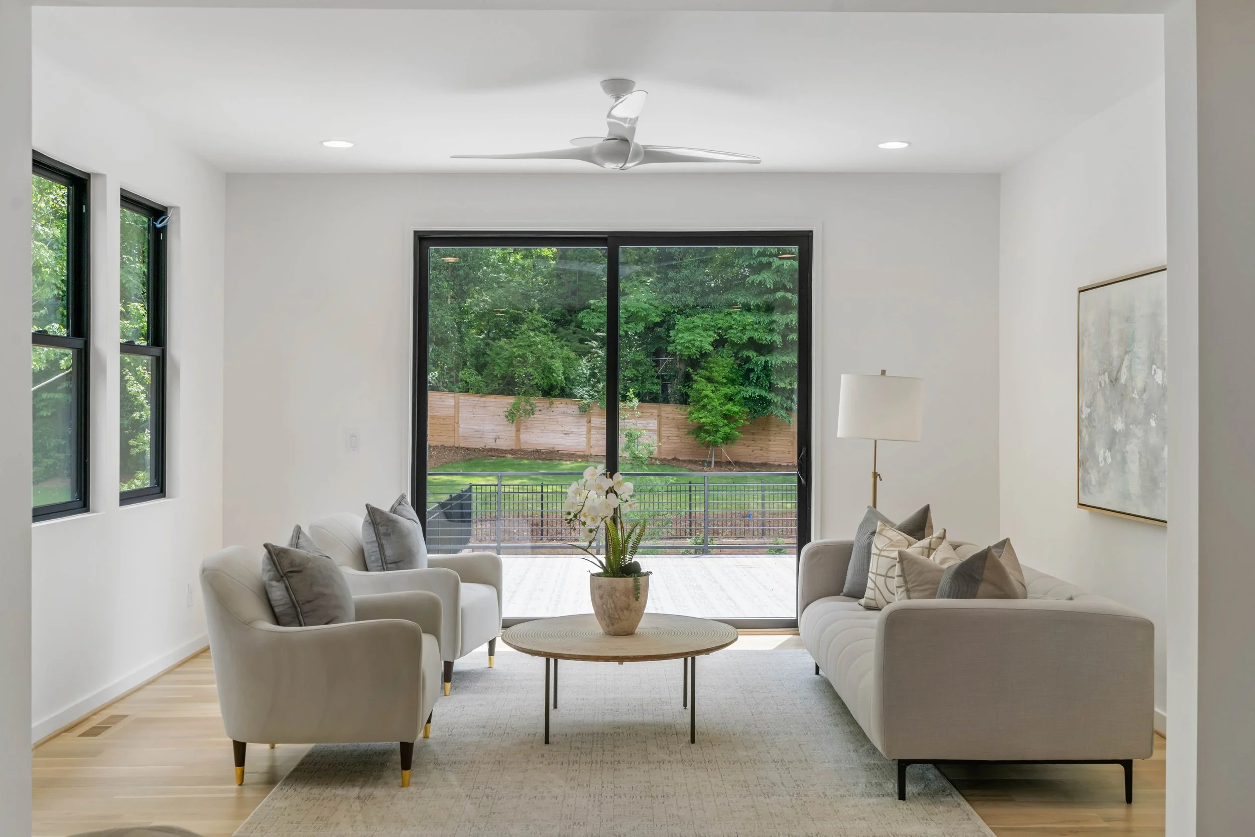 Modern living room with white sofas and gray cushions, a wooden coffee table with a potted orchid, large sliding glass door leading to a backyard, minimalistic wall art, white walls, hardwood flooring, and a ceiling fan.