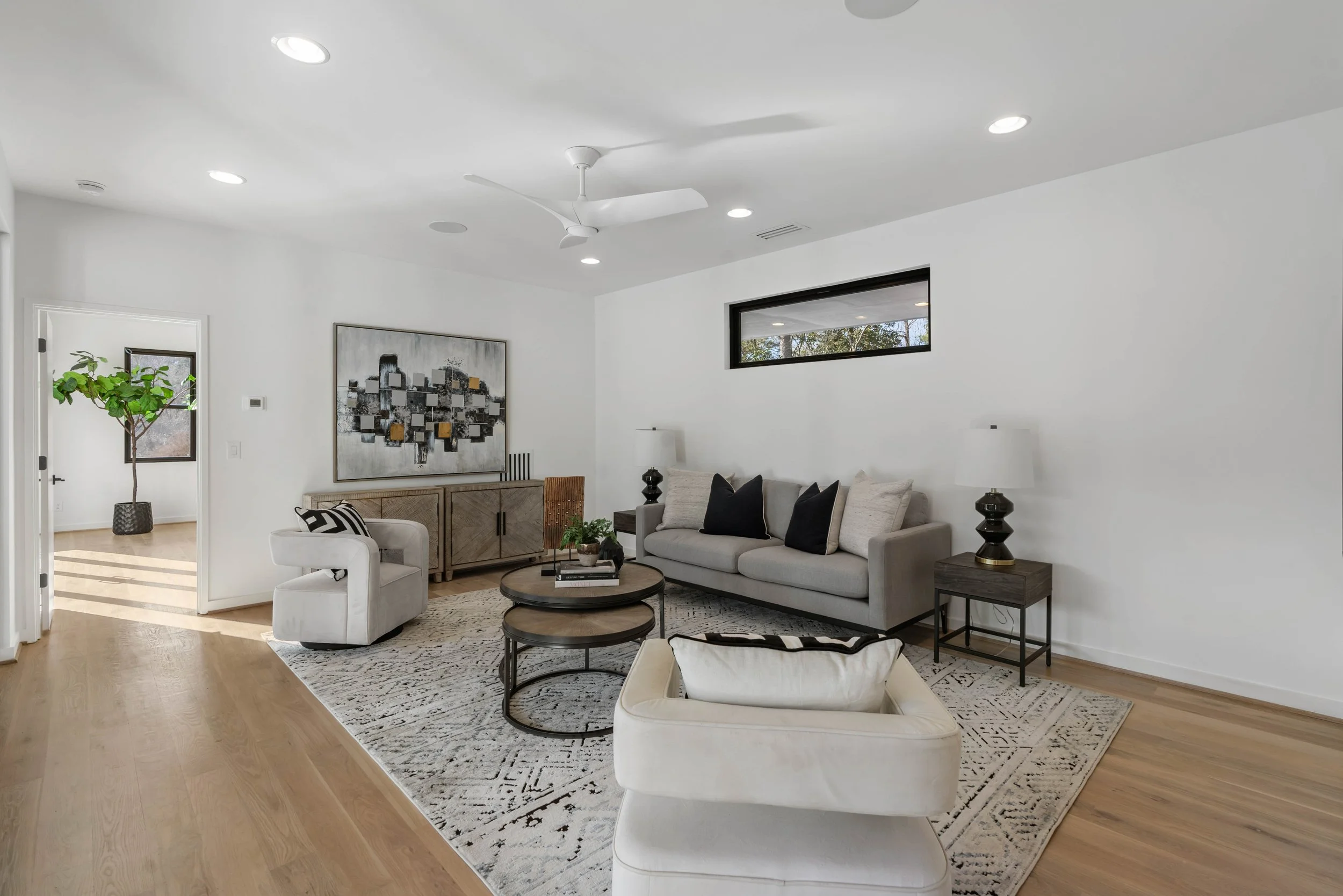 Modern living room with gray sofa, two white armchairs, a black and gray rug, a light wood floor, black side tables with lamps, a wooden cabinet, abstract wall art, and a small window high on the wall.