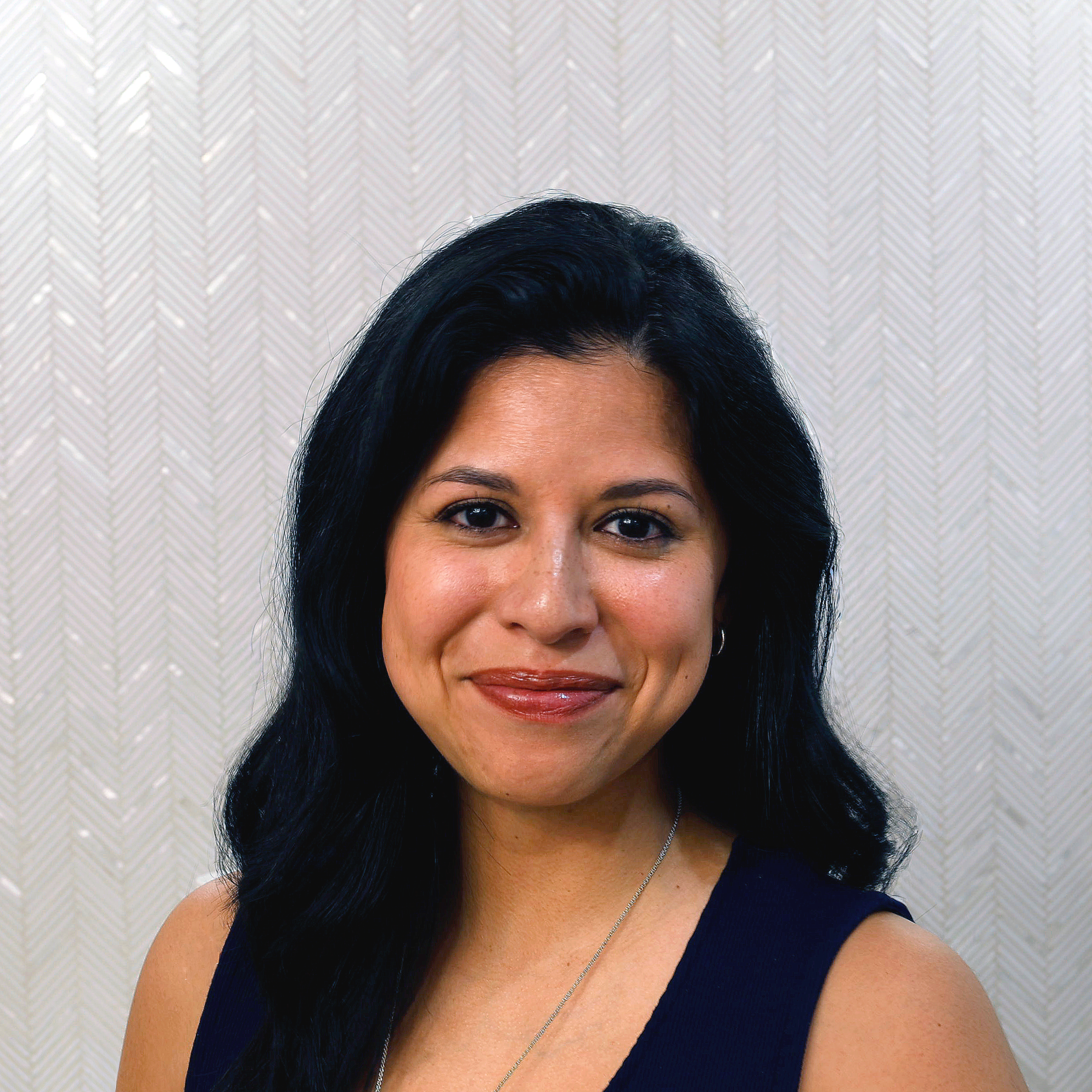 A smiling woman with long black hair wearing a black sleeveless top, a gold necklace, and earrings, standing in front of a light-colored, textured background.
