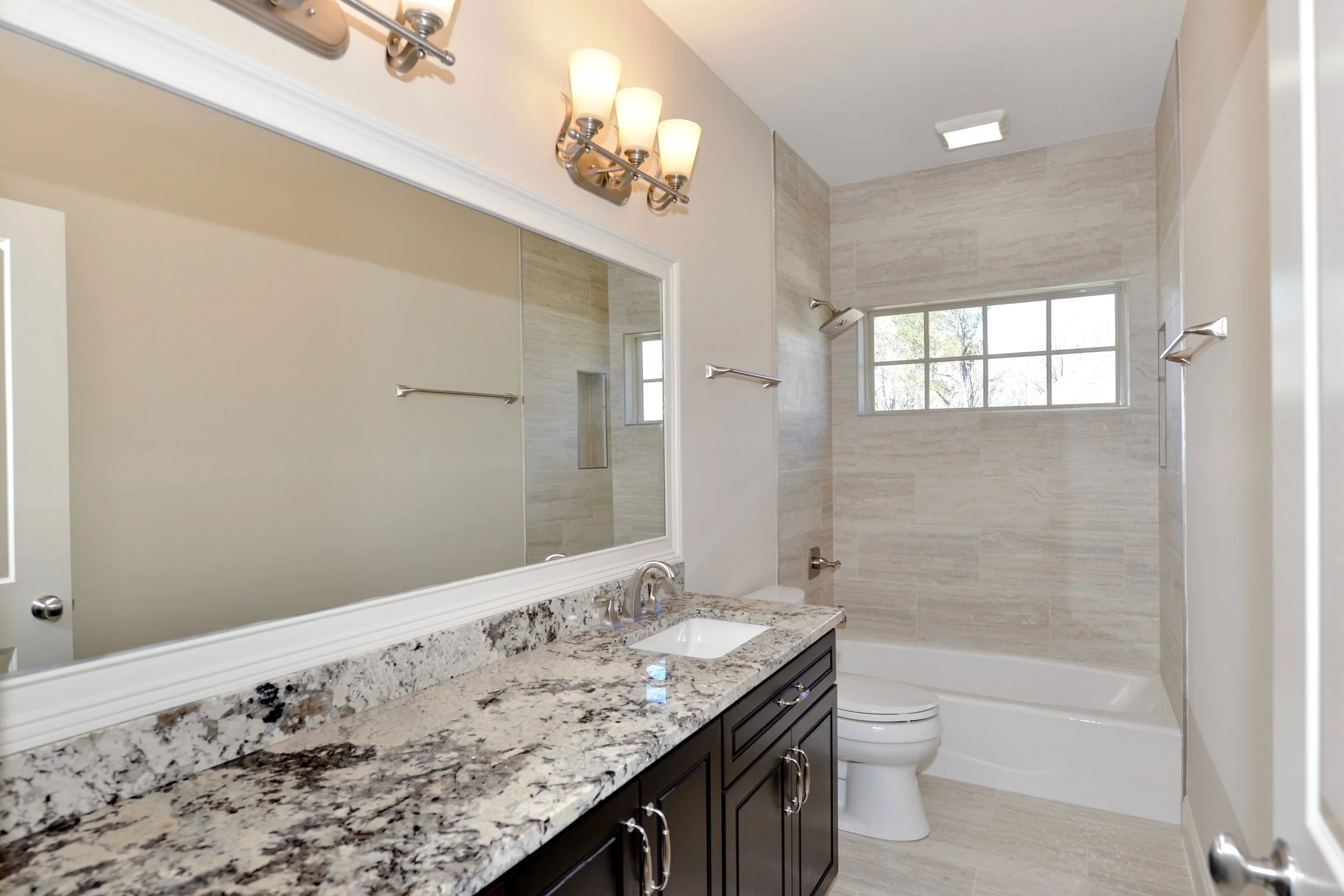 Modern bathroom with granite countertop vanity, large mirror, wall-mounted light fixture, shower area with window, and beige tile walls.