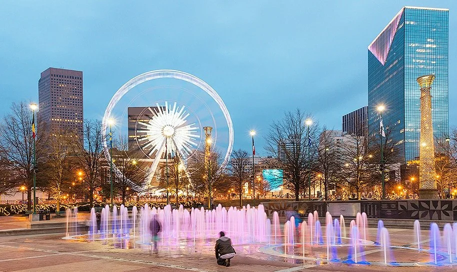 Colorful fountains in a city park with a Ferris wheel, skyscrapers, and glowing lights in the background at dusk.