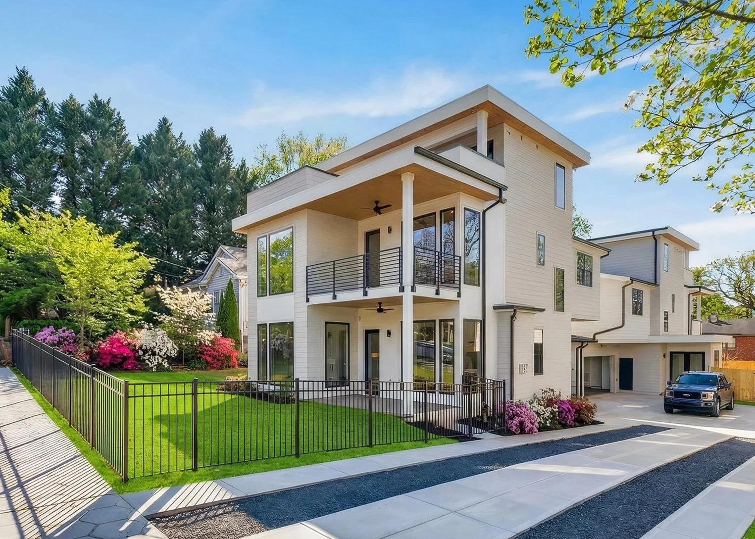 Modern two-story house with large windows, balconies, a fenced yard, blooming flowers, and a driveway under a clear blue sky.