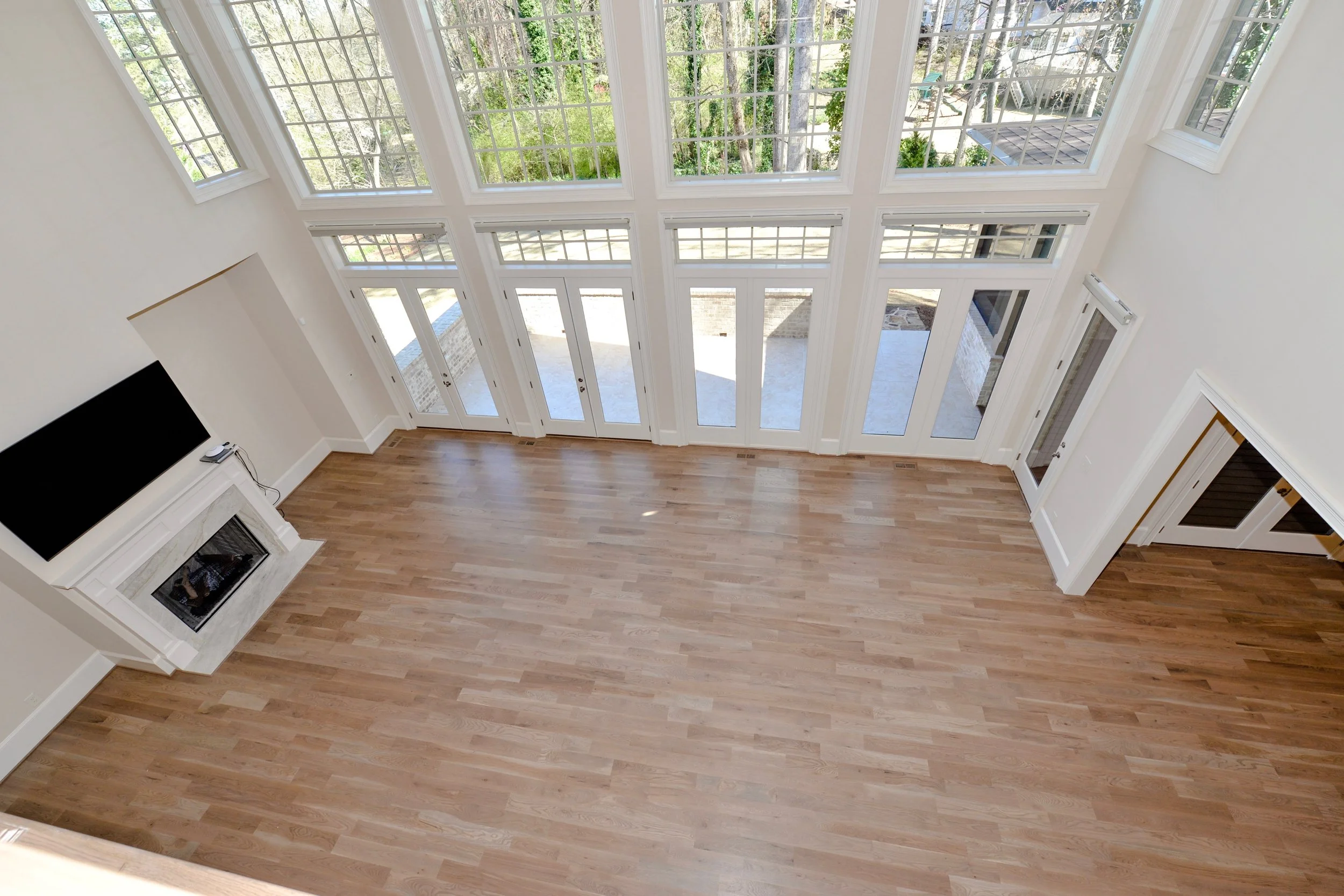 Empty living room with hardwood floors, large windows, and a fireplace with a mounted TV above it.